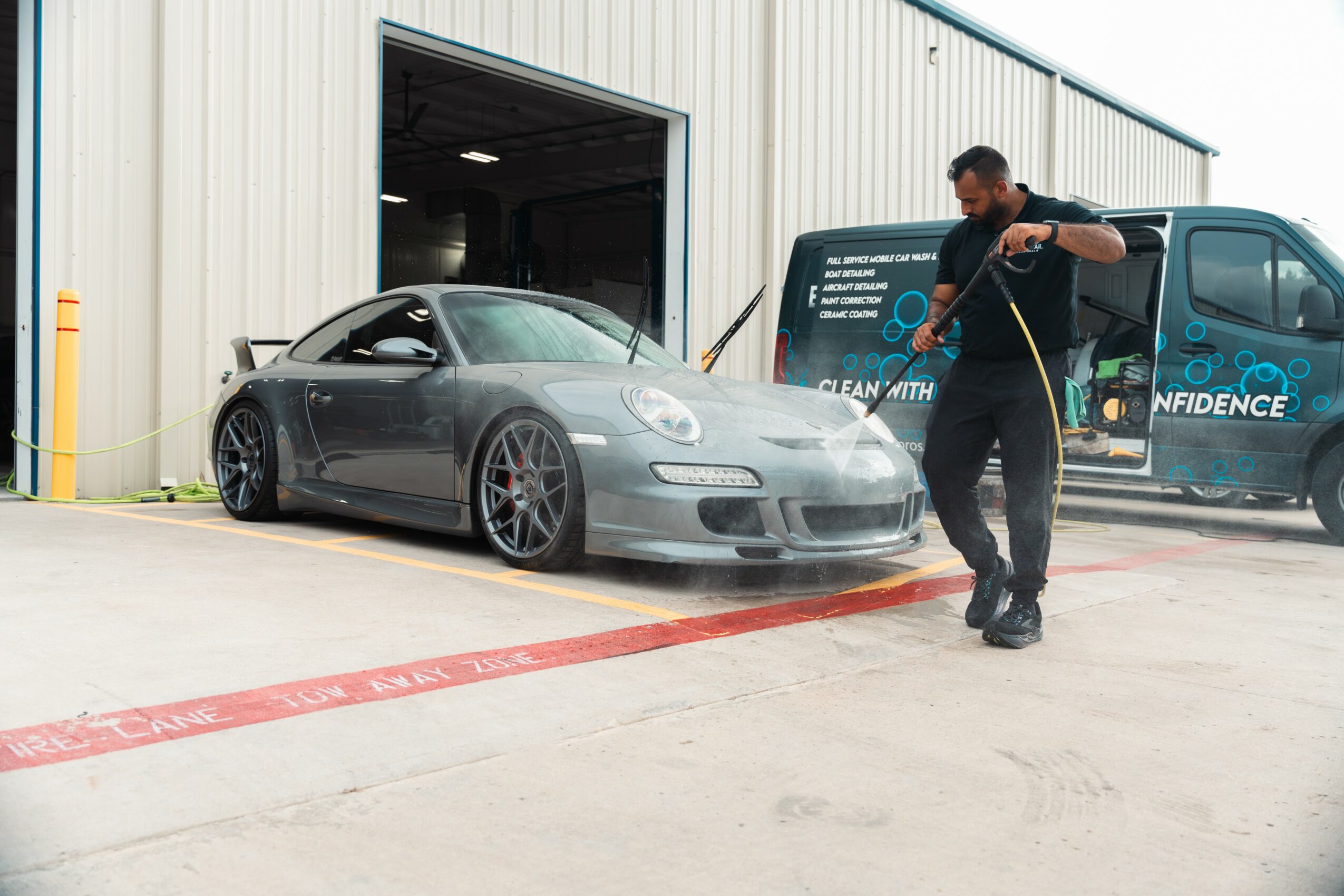 Person cleaning a gray sports car outside a large industrial building with a van in the background.