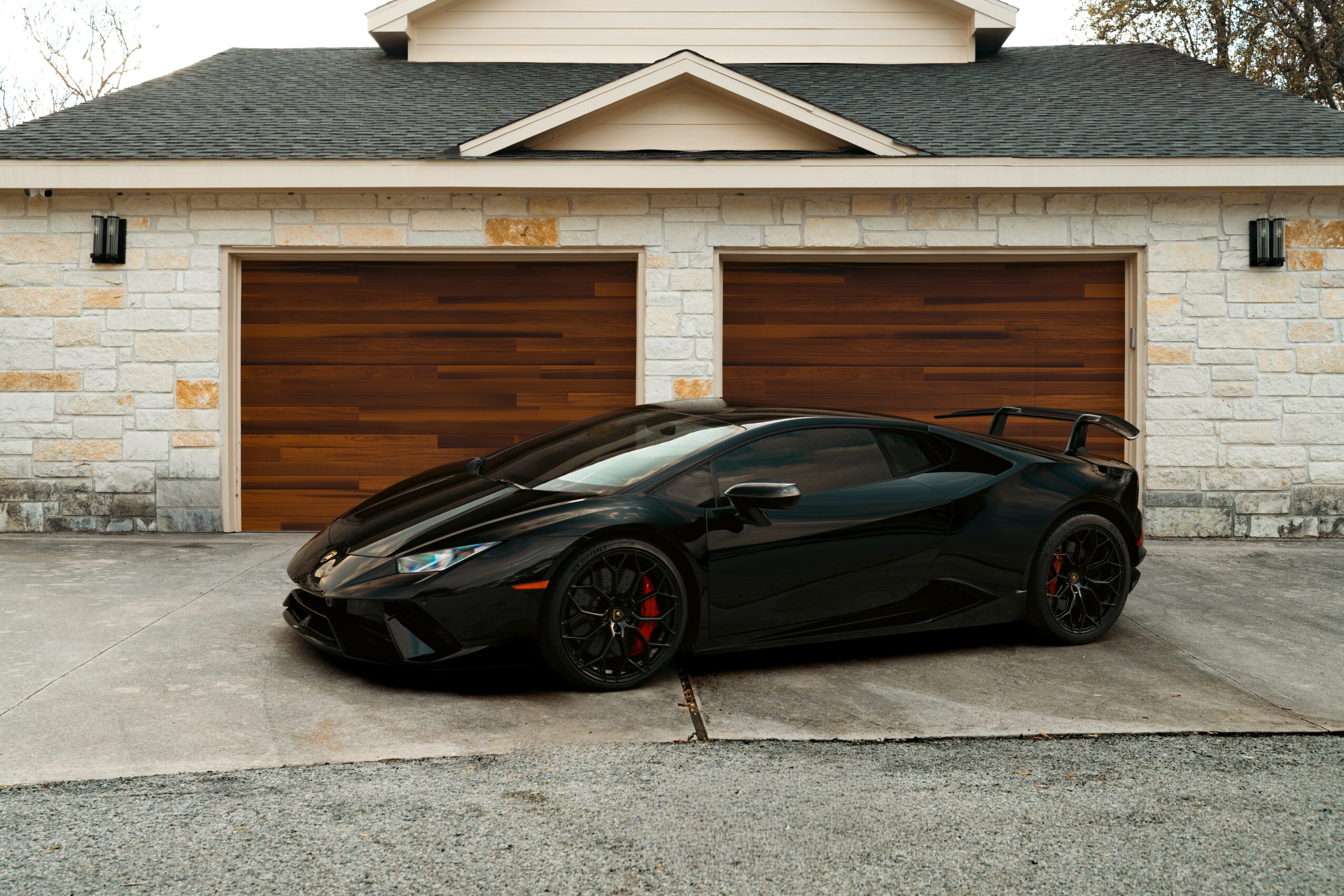 Black sports car parked in front of a garage with two wooden doors, stone exterior, and a small roof overhang