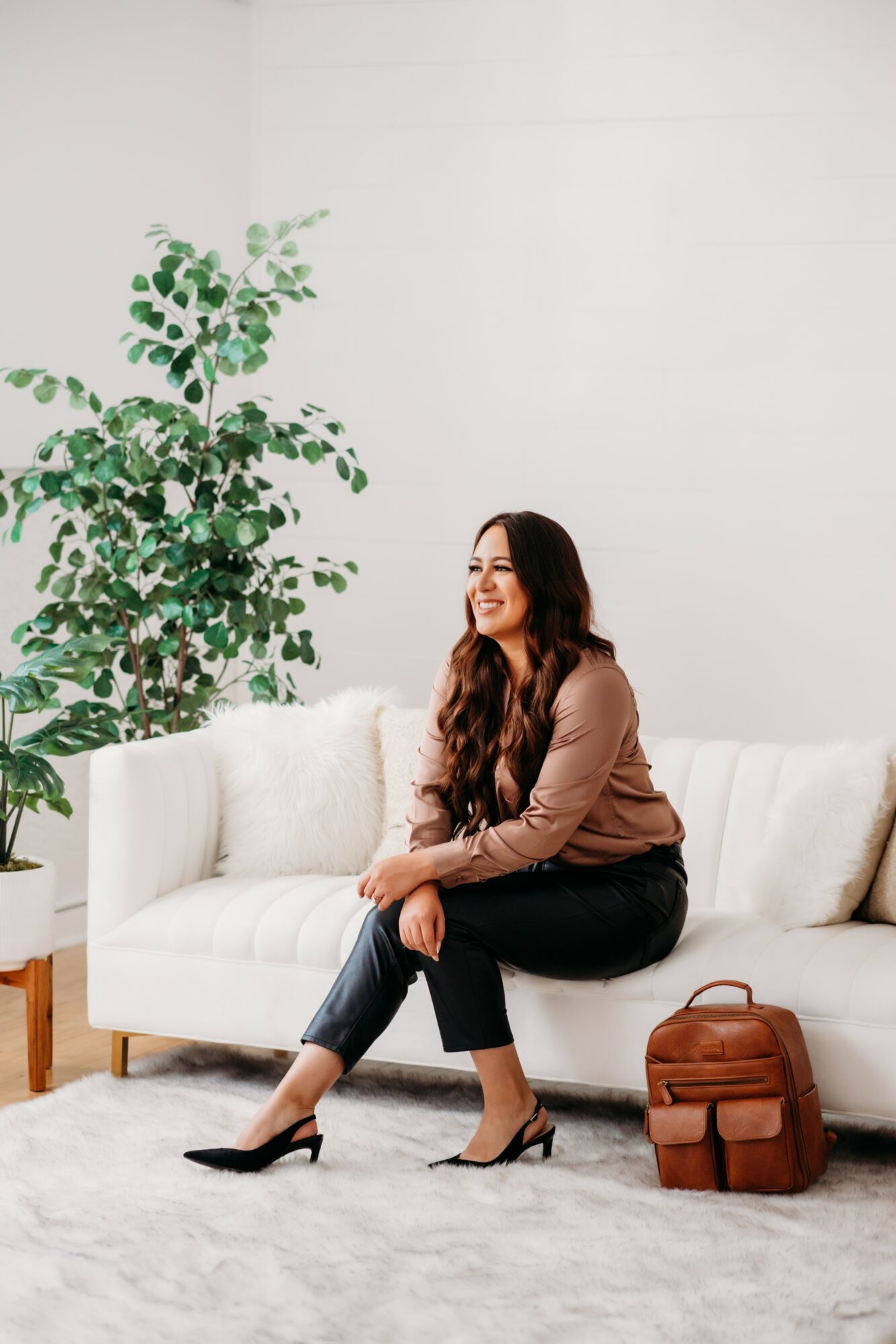 Woman with long hair sitting on a white sofa, smiling, in a bright room with a large plant, a brown bag, and a white rug.