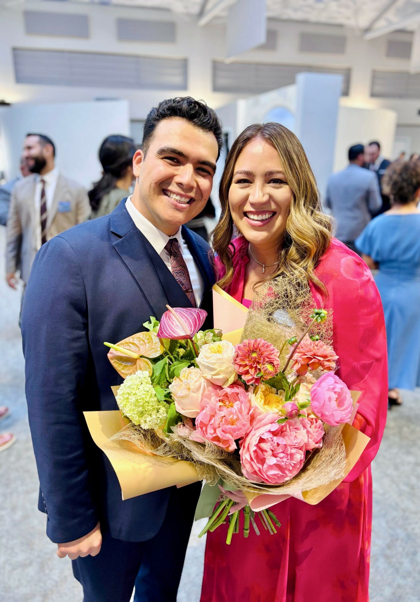 Two smiling people, one holding a large bouquet of pink and white flowers, at a formal event with others in background.