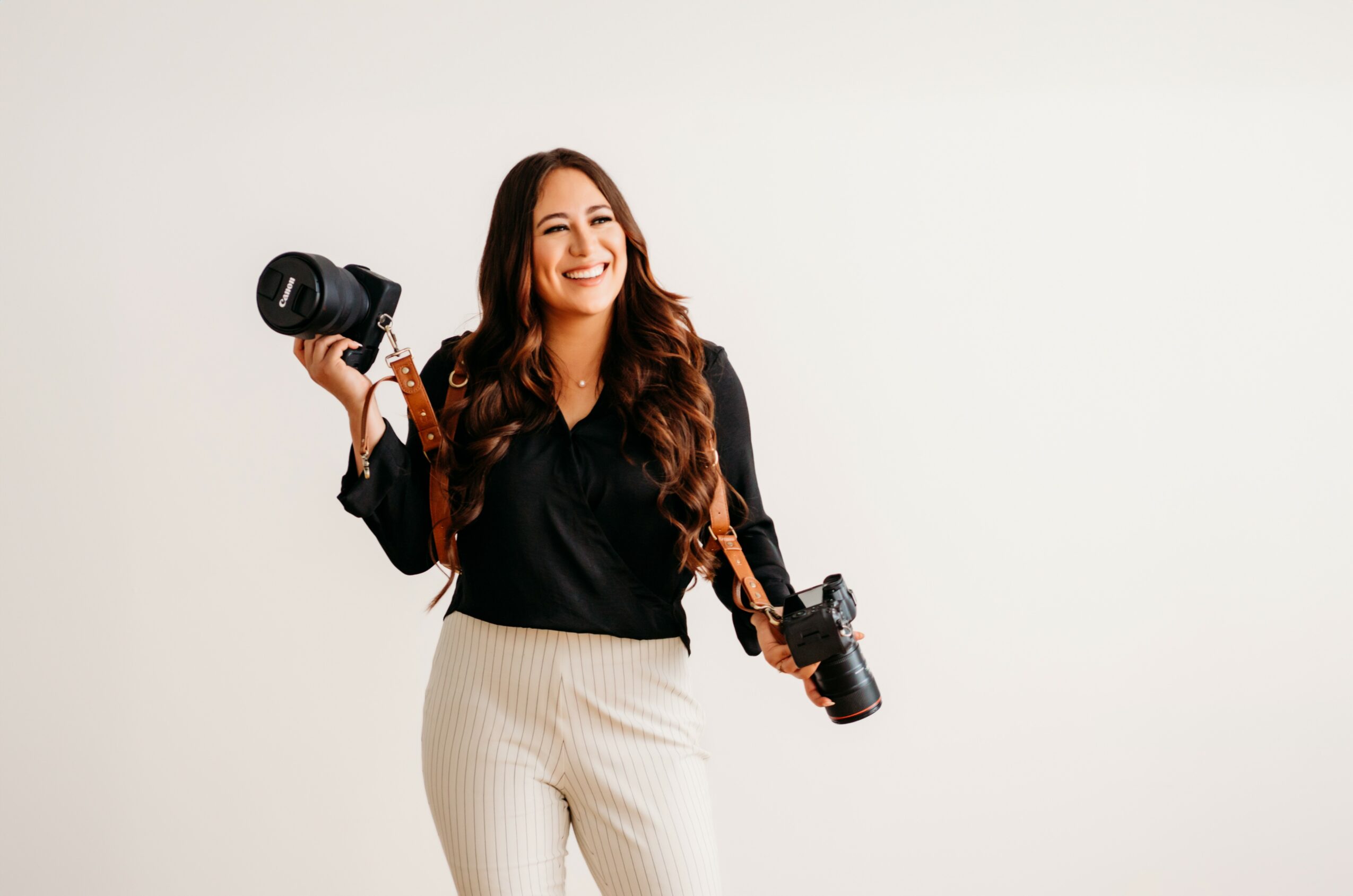 Woman with long hair holding two cameras, smiling, against a plain white background.