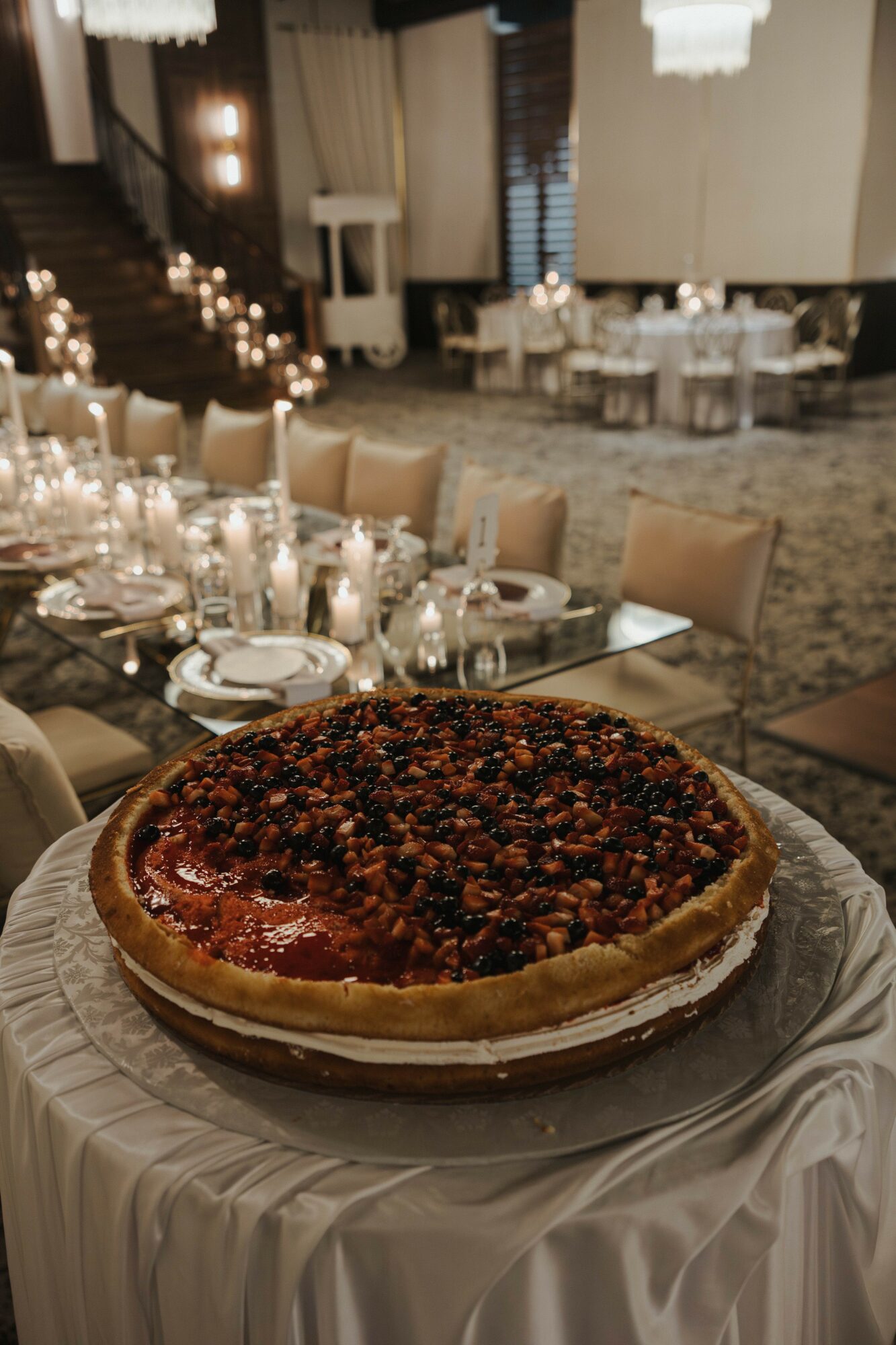 A large berry tart on a table in an elegant banquet hall with decorated tables and chairs, dim lighting, and chandeliers.