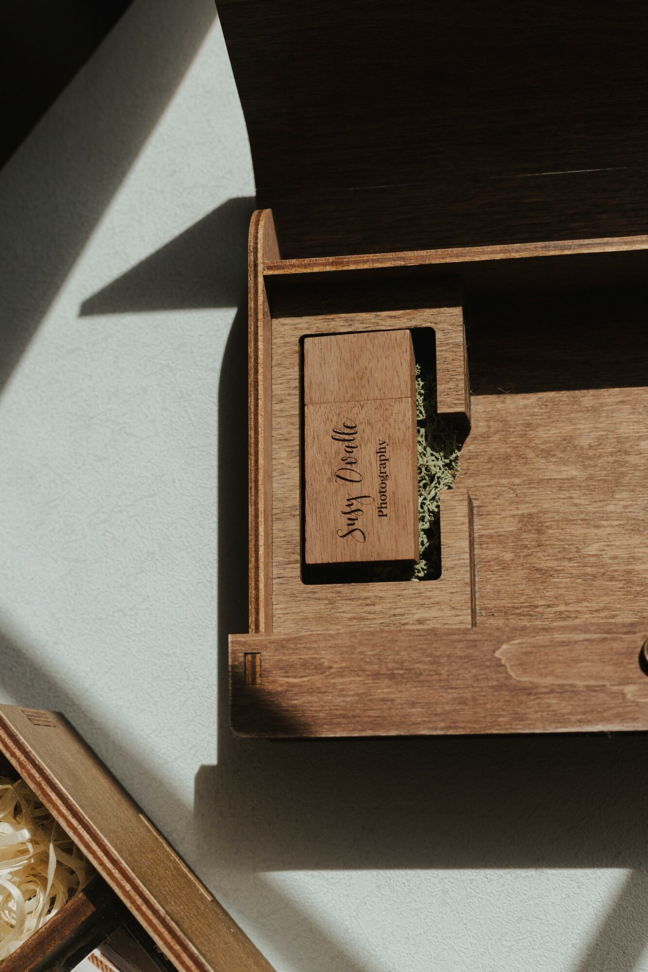 Open wooden box with a small compartment containing a wooden plaque and some dried moss, on a surface with shadows.