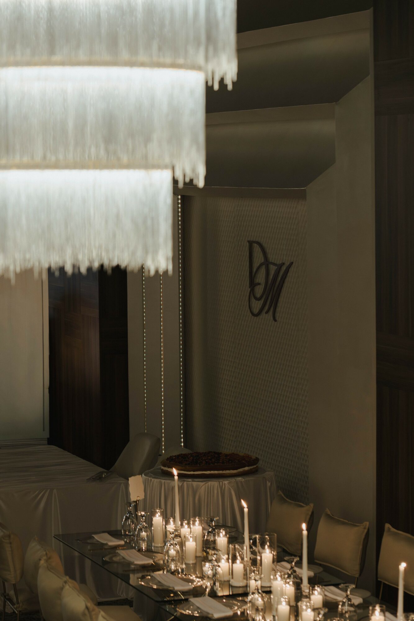Part of a dining area with a chandelier, table set with candles, and a wall with decorative initials.