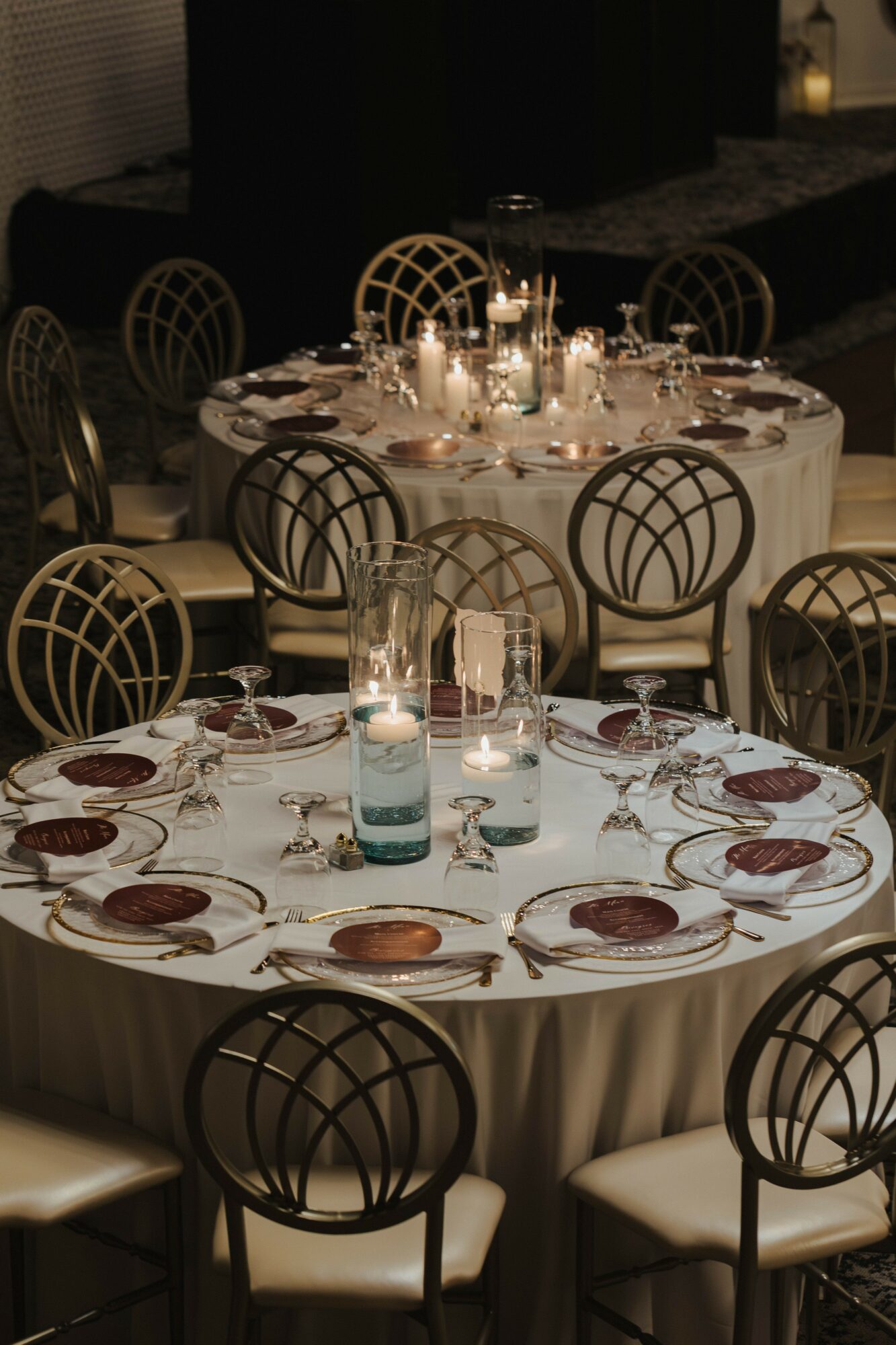 Two round tables set for a formal event with candles, glasses, and plates, surrounded by elegant chairs, dimly lit background.