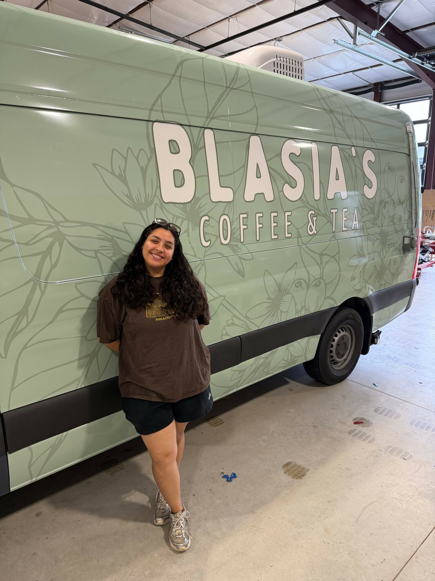 Woman standing next to a large van with 'BLASV Coffee' logo, indoors, smiling, wearing a dark jacket and shorts.