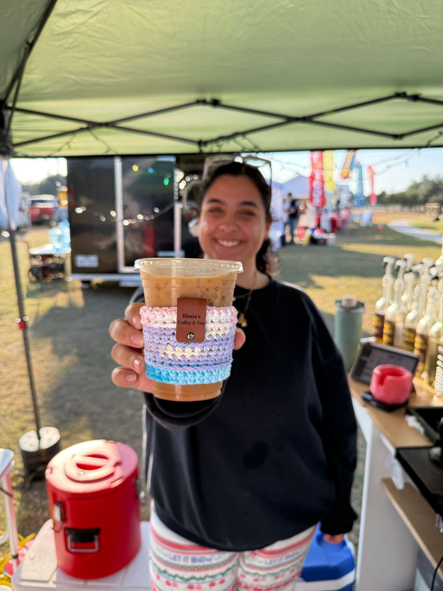 Woman holding a colorful layered cake with a small figure on top, outdoors at a fair or amusement park.