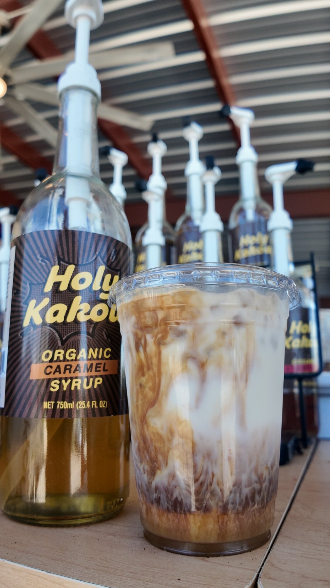A plastic cup with iced coffee and swirls of milk next to a bottle labeled Holy Kakao, organic caramel syrup, on a wooden surface.