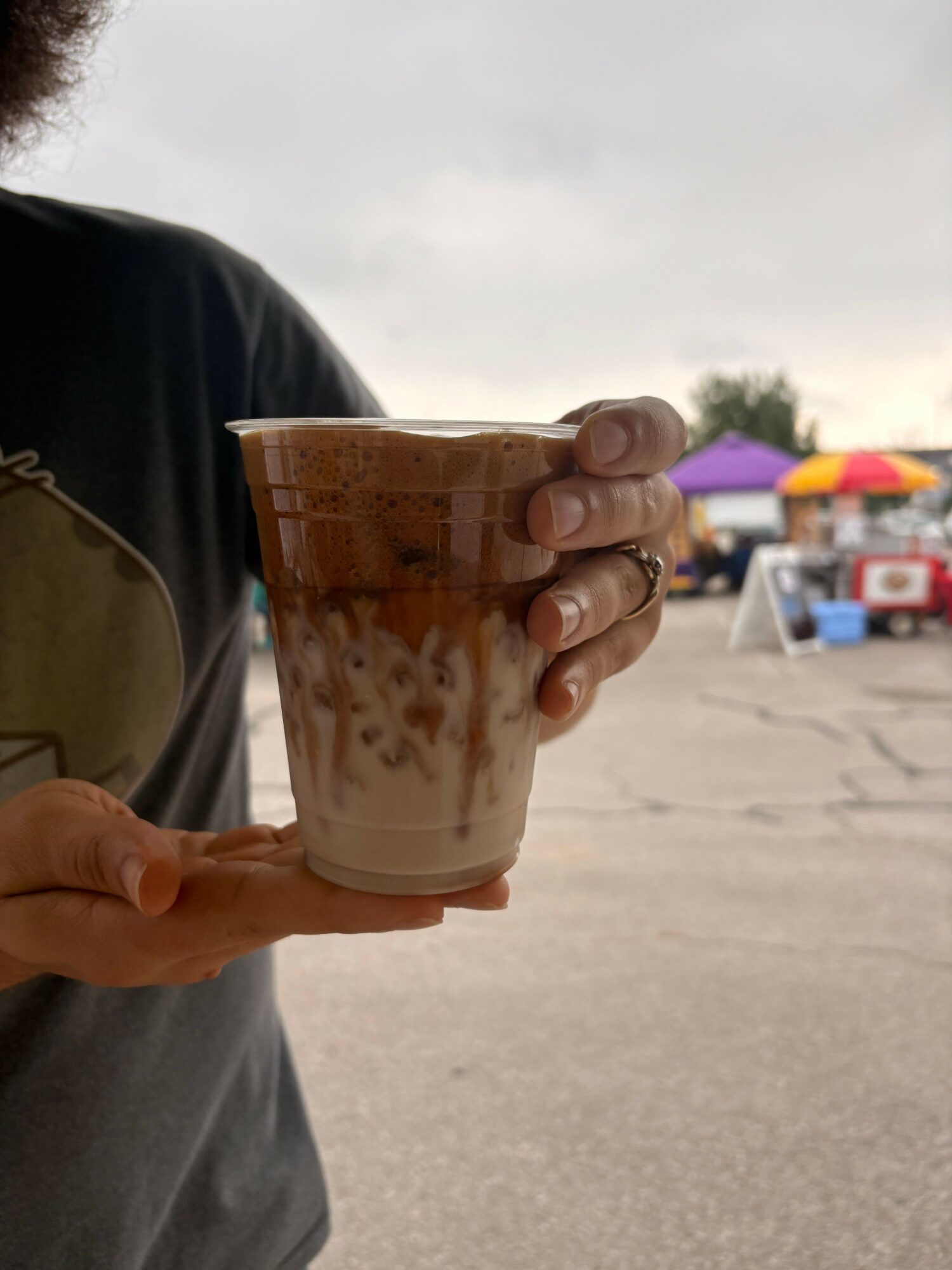Person holding a plastic cup with a beverage, outdoors on a cloudy day, with colorful tents in the background.