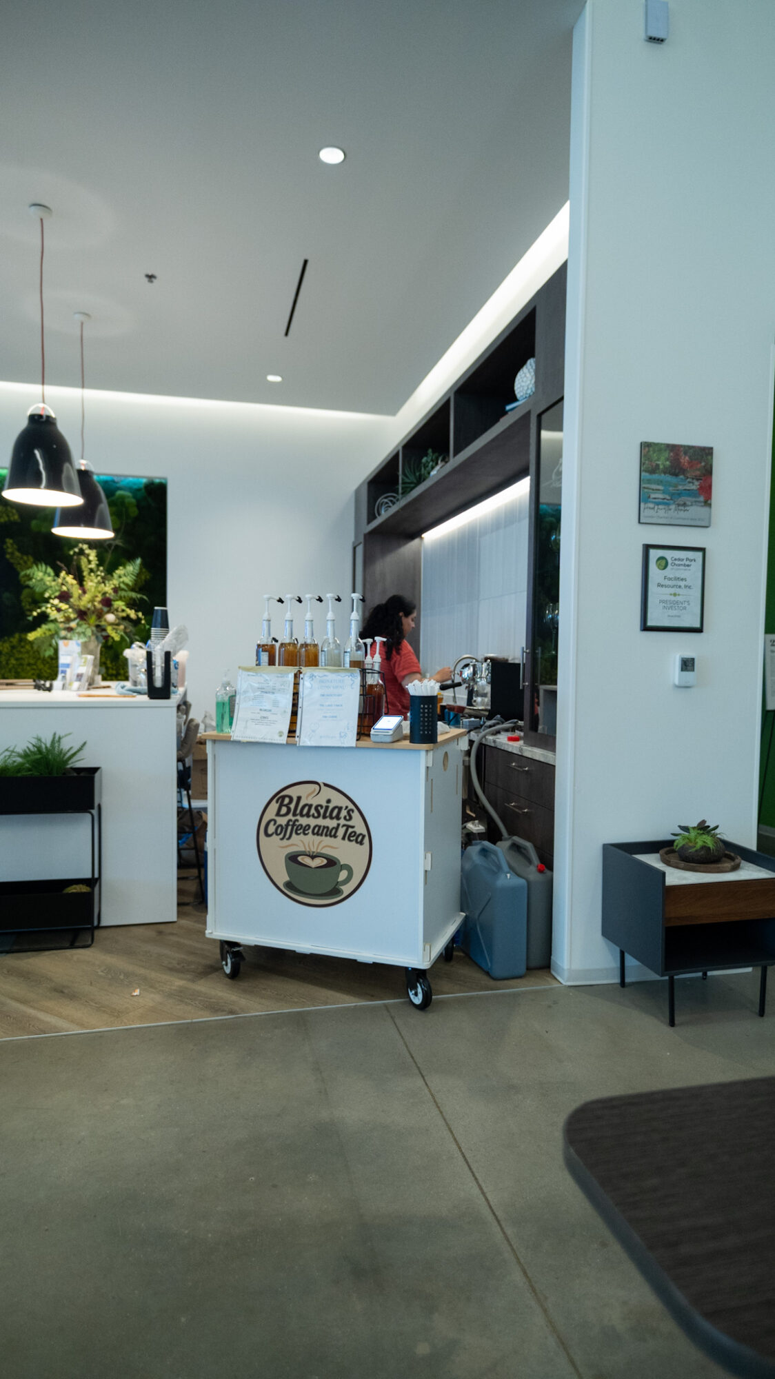 Interior of a cafe with a counter, shelves, and a person working behind the counter, bright lighting, and modern decor.