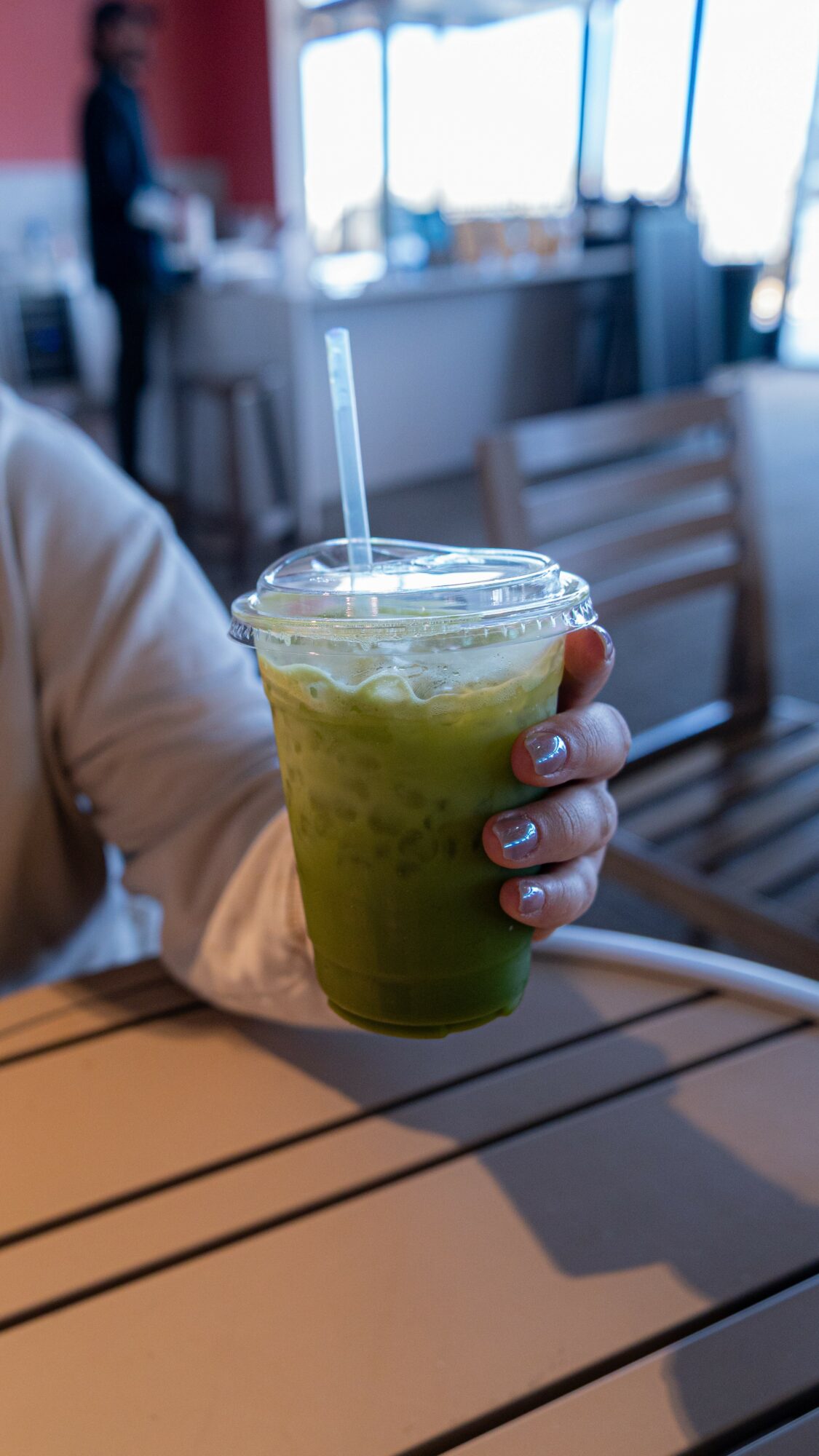 Person holding a green iced beverage with a straw in a cafe setting, with tables and chairs visible in the background.