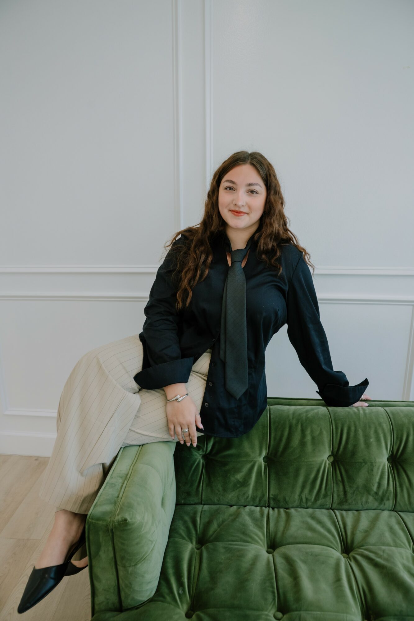 Young woman with long curly hair leaning on a green velvet sofa, smiling, in a light-colored room.