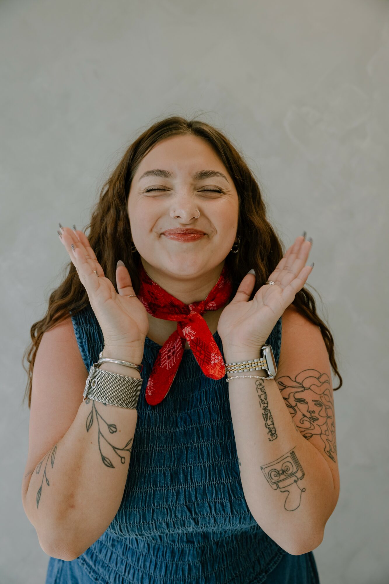 Young woman with long hair, smiling with eyes closed, wearing a red bandana and blue top, holding her face with hands.