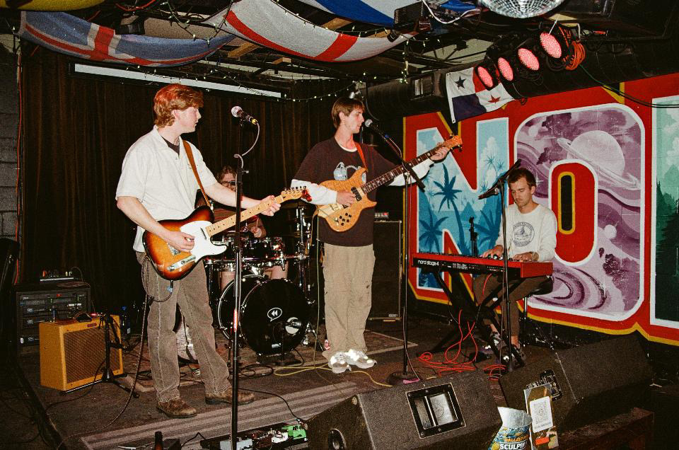 Band performing on stage with three musicians playing guitar, keyboard, and drums, and a colorful mural background.