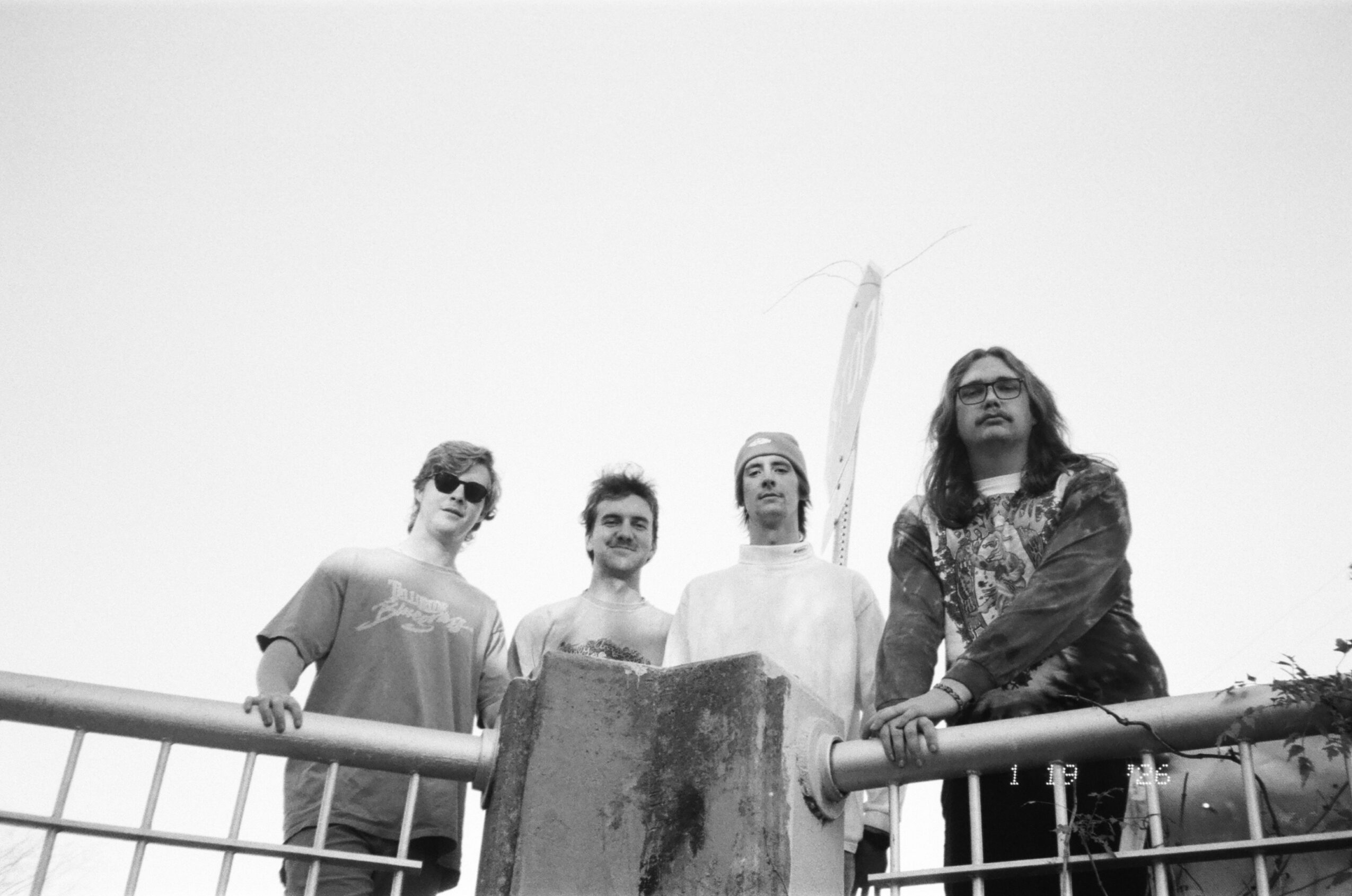 Four people stand behind a railing, looking at the camera, with a plain sky background.