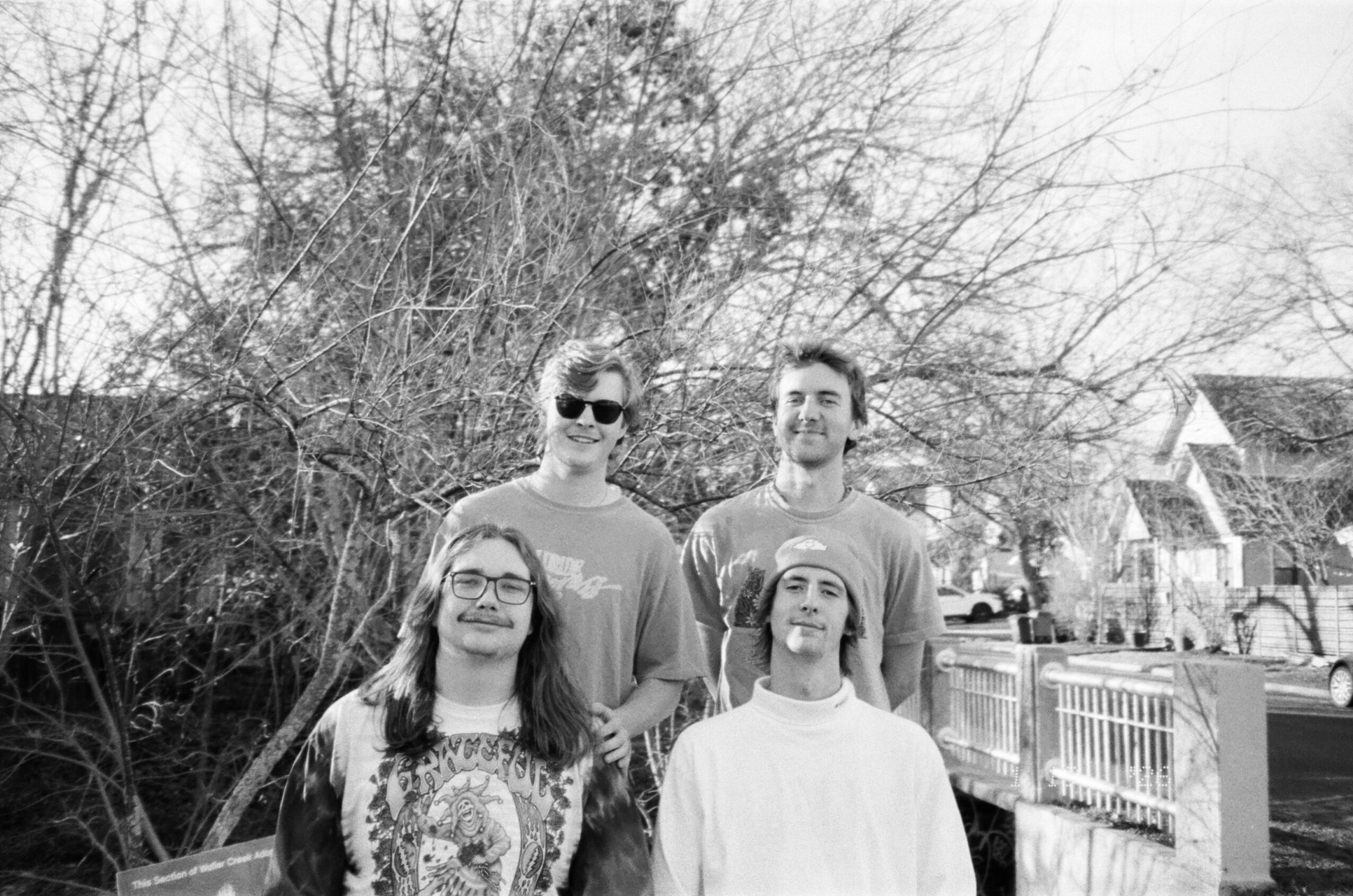 Four people standing outdoors in front of trees and houses, some wearing glasses, in black and white.