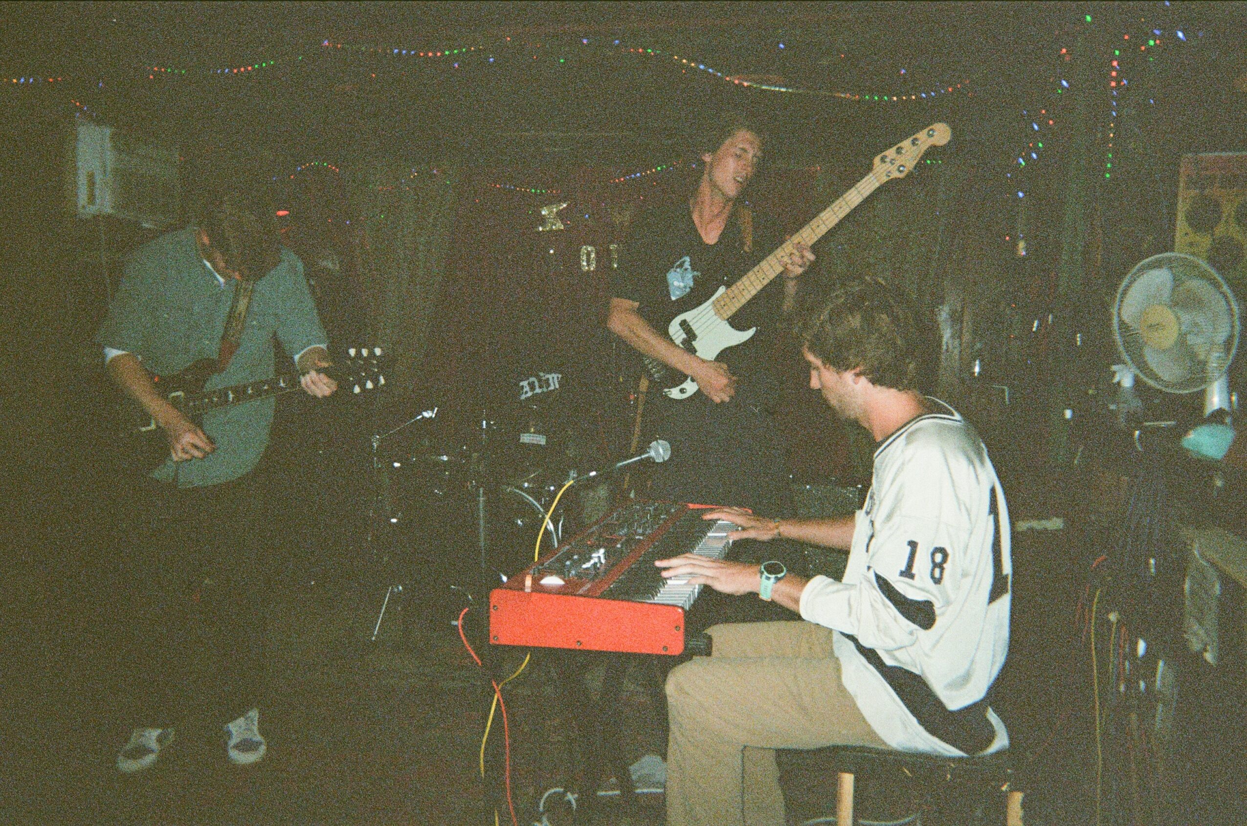 Three musicians perform on stage with guitars and keyboard, in a dimly lit setting.