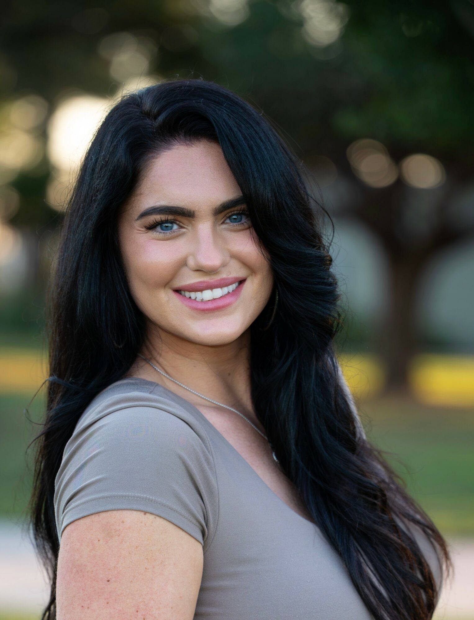 Young woman with long dark hair smiling outdoors, wearing a gray top, with trees in the background.