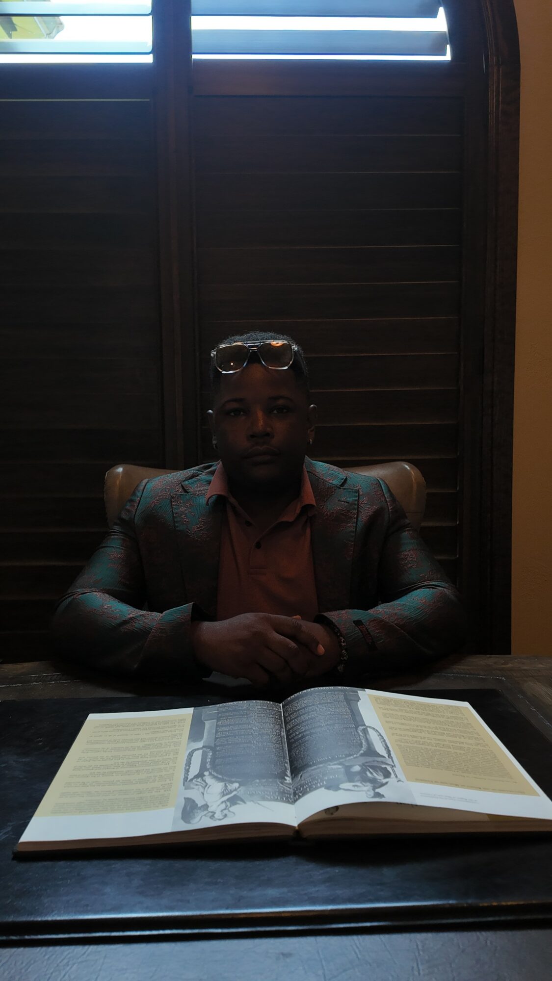 Person sitting at a table with an open book, wearing glasses and a suit, in a dimly lit room.