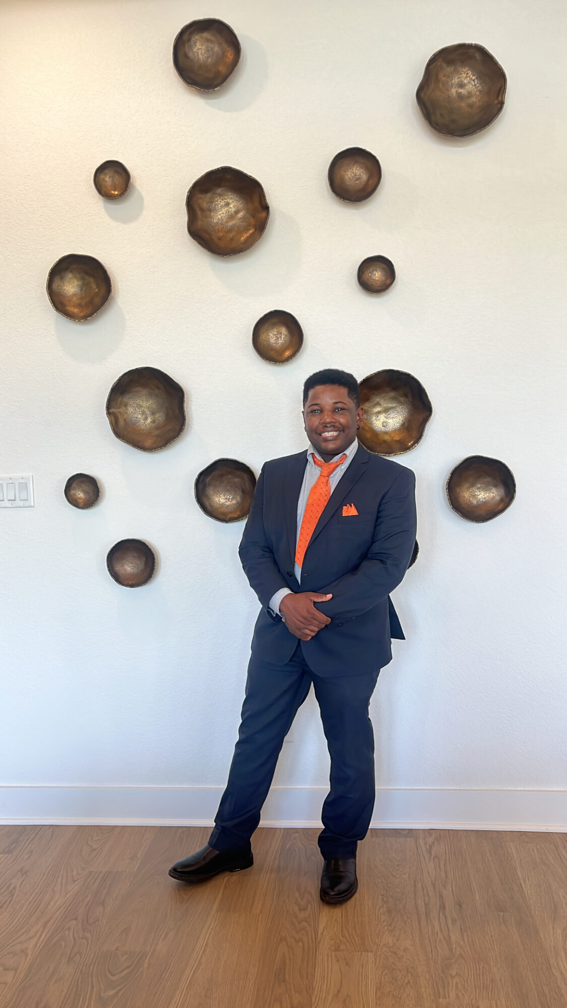 Man in a suit with an orange tie standing in front of a wall with round decorative objects.