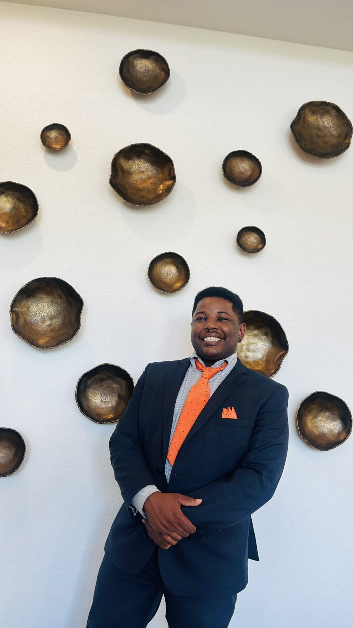 Man in a suit with an orange tie and pocket square, smiling, standing in front of a white wall with round metallic art pieces.
