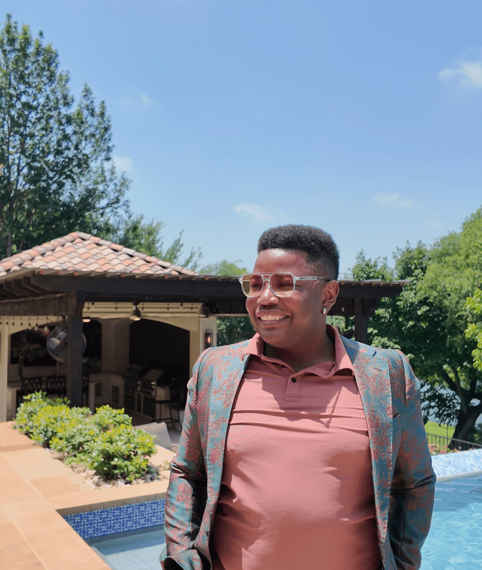 Young man with glasses smiling outdoors, wearing a pink shirt and a patterned blazer, standing near a pool with trees and a building in background.
