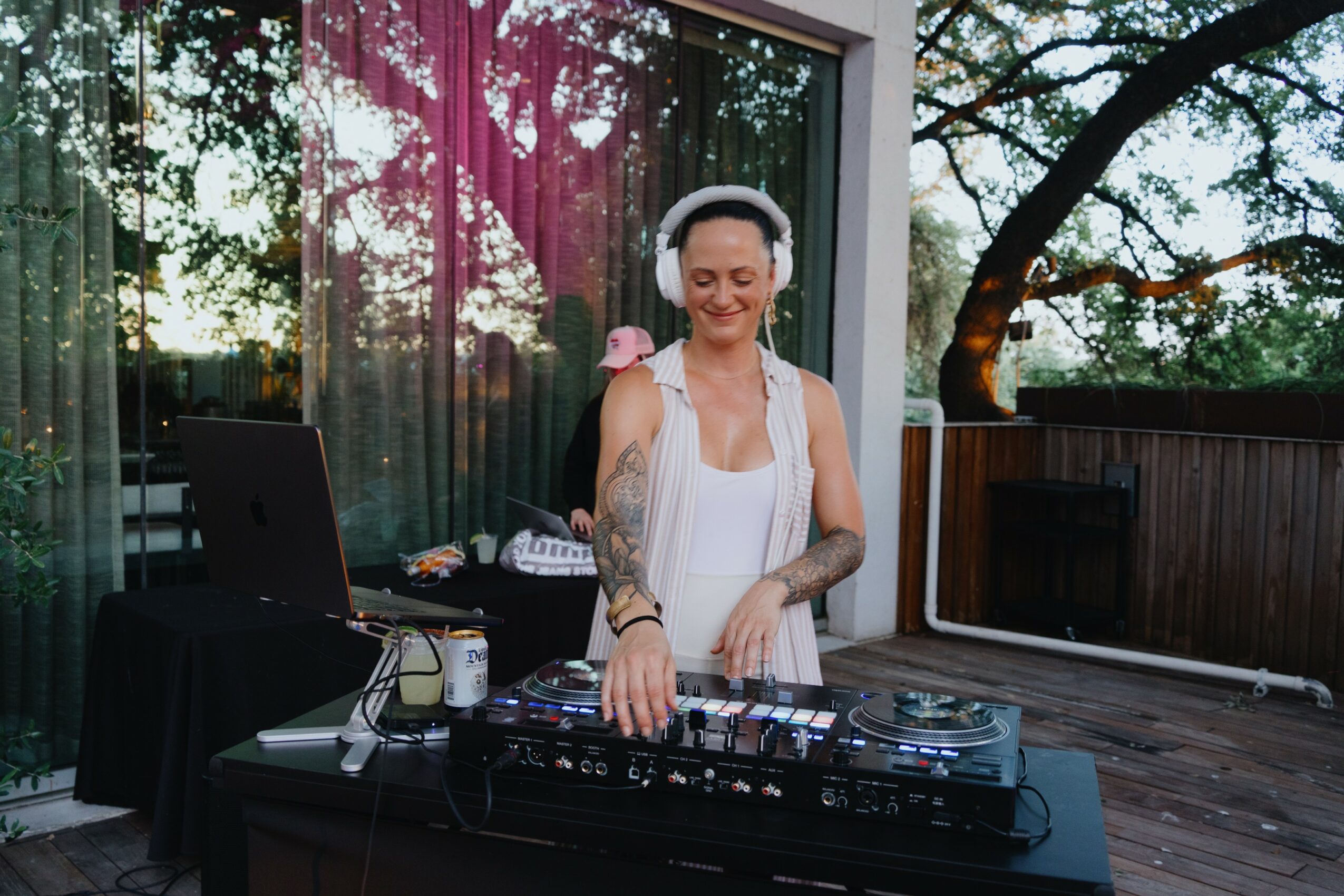 Woman DJ wearing headphones, standing outdoors with trees and a wooden deck, using DJ equipment on a table.