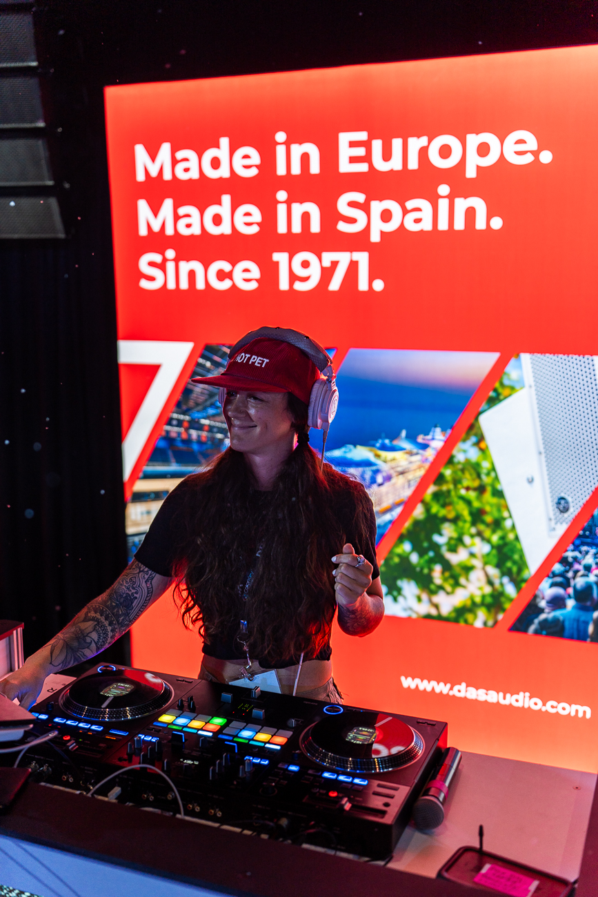 Woman with headphones and a cap standing behind DJ equipment, smiling, in front of a red and white promotional display.