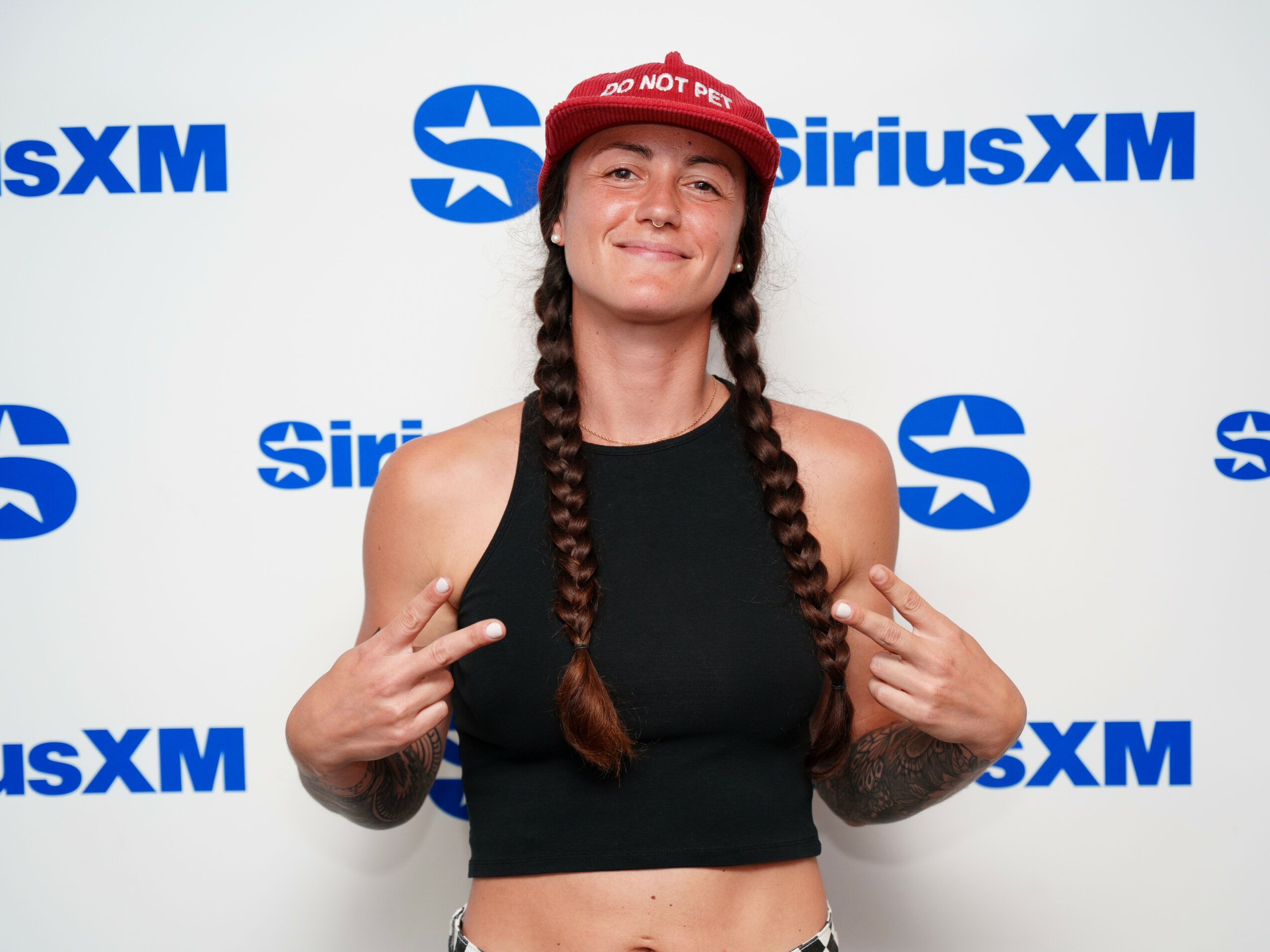 Woman with long braided hair wearing a red cap and black sleeveless top, smiling, in front of SiriusXM backdrop.