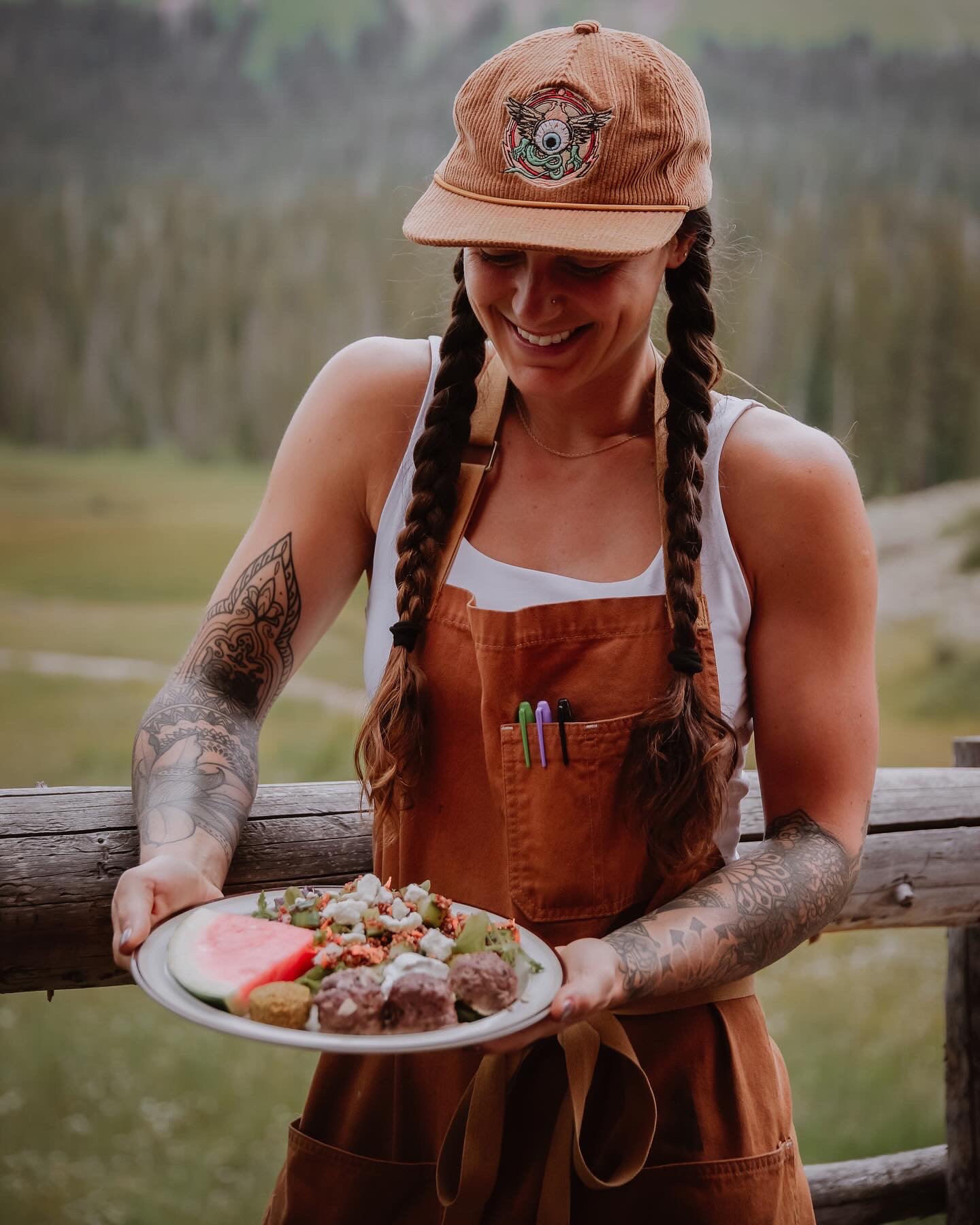 Woman with braided hair, wearing a cap and apron, holding a plate of food outdoors, smiling.