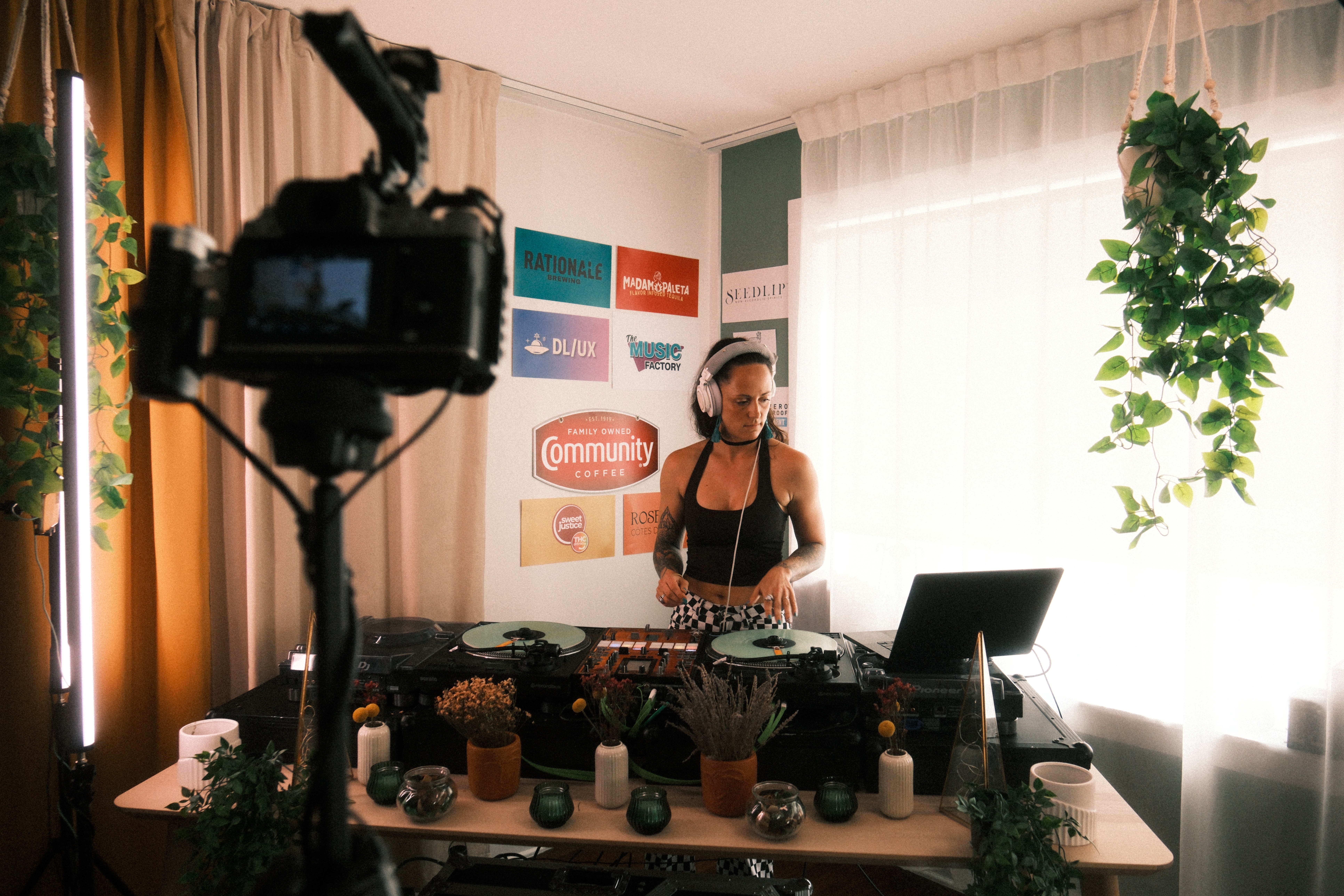 Woman with headphones standing behind a DJ setup in a room with natural light, plants, and colorful signs on the wall.