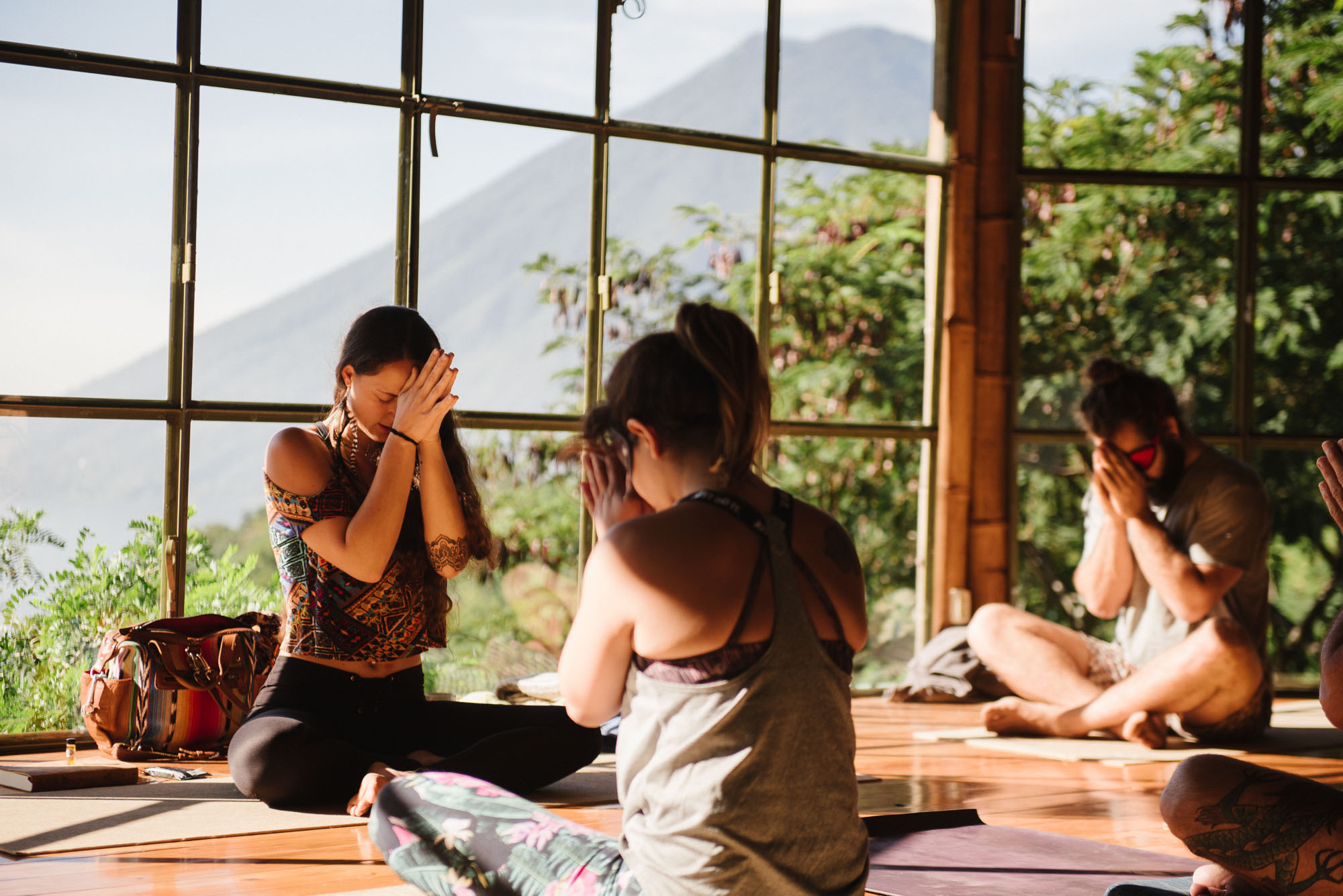 Three women practicing yoga or meditation indoors near large windows with a mountain view, sitting cross-legged with hands in prayer position.
