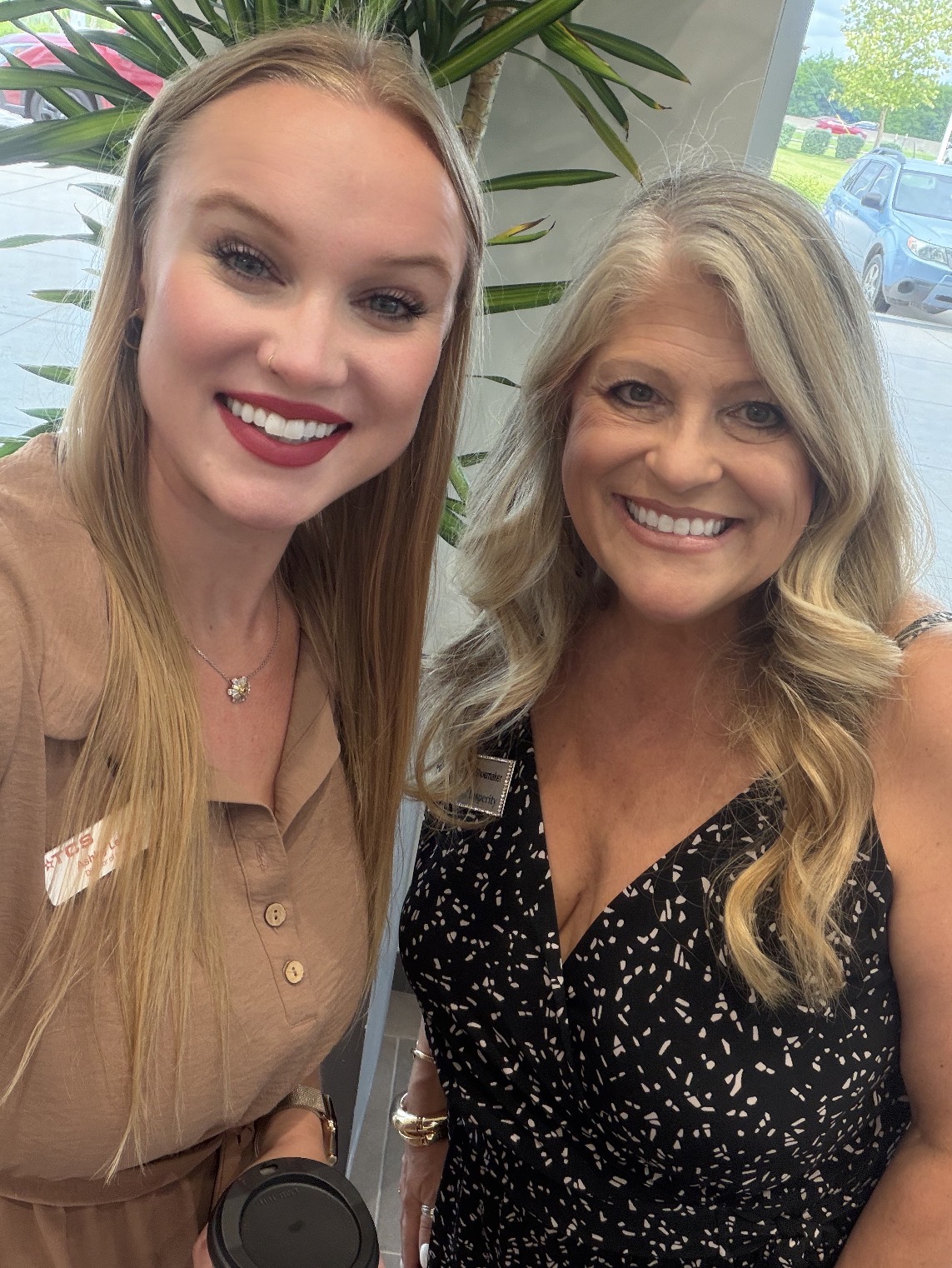 Two women smiling indoors with plants and a window in the background.