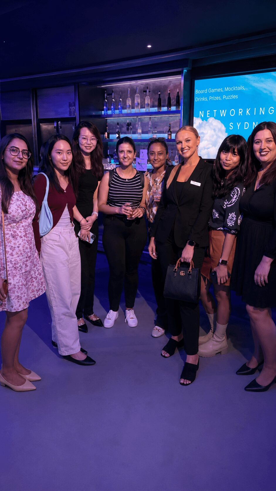 Group of seven women standing together indoors, some holding drinks, with a bar and a screen in the background.