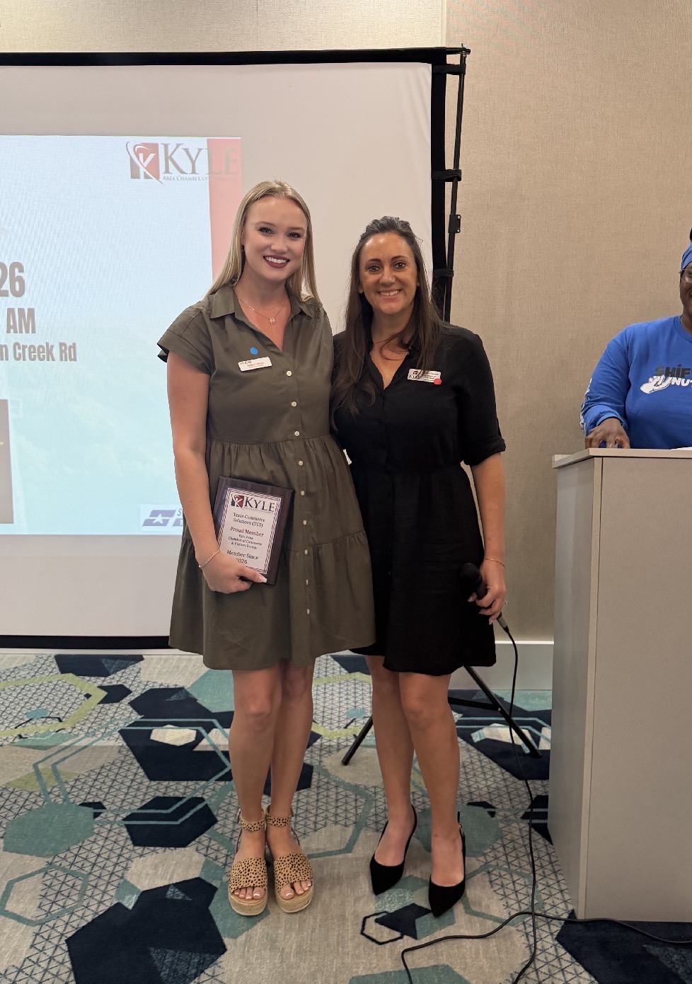 Two women standing side by side in front of a presentation screen, smiling, one holding a certificate, in a conference room.