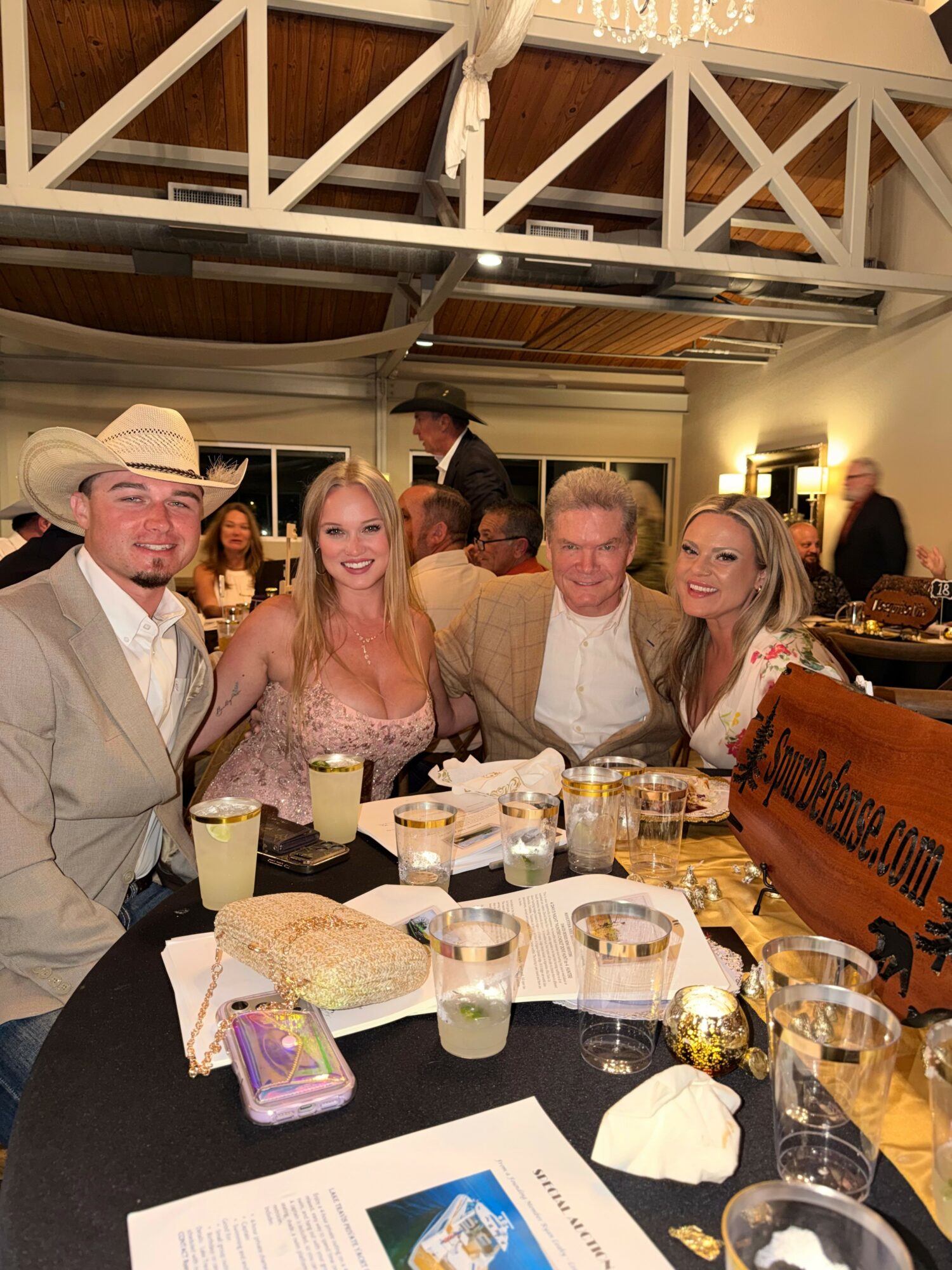 Four people in formal attire sitting at a table with drinks and food, smiling at a social event indoors.