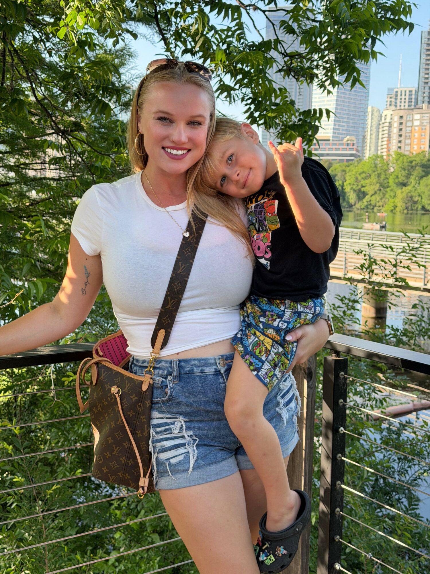 A woman holding a child outdoors with trees and city buildings in the background.