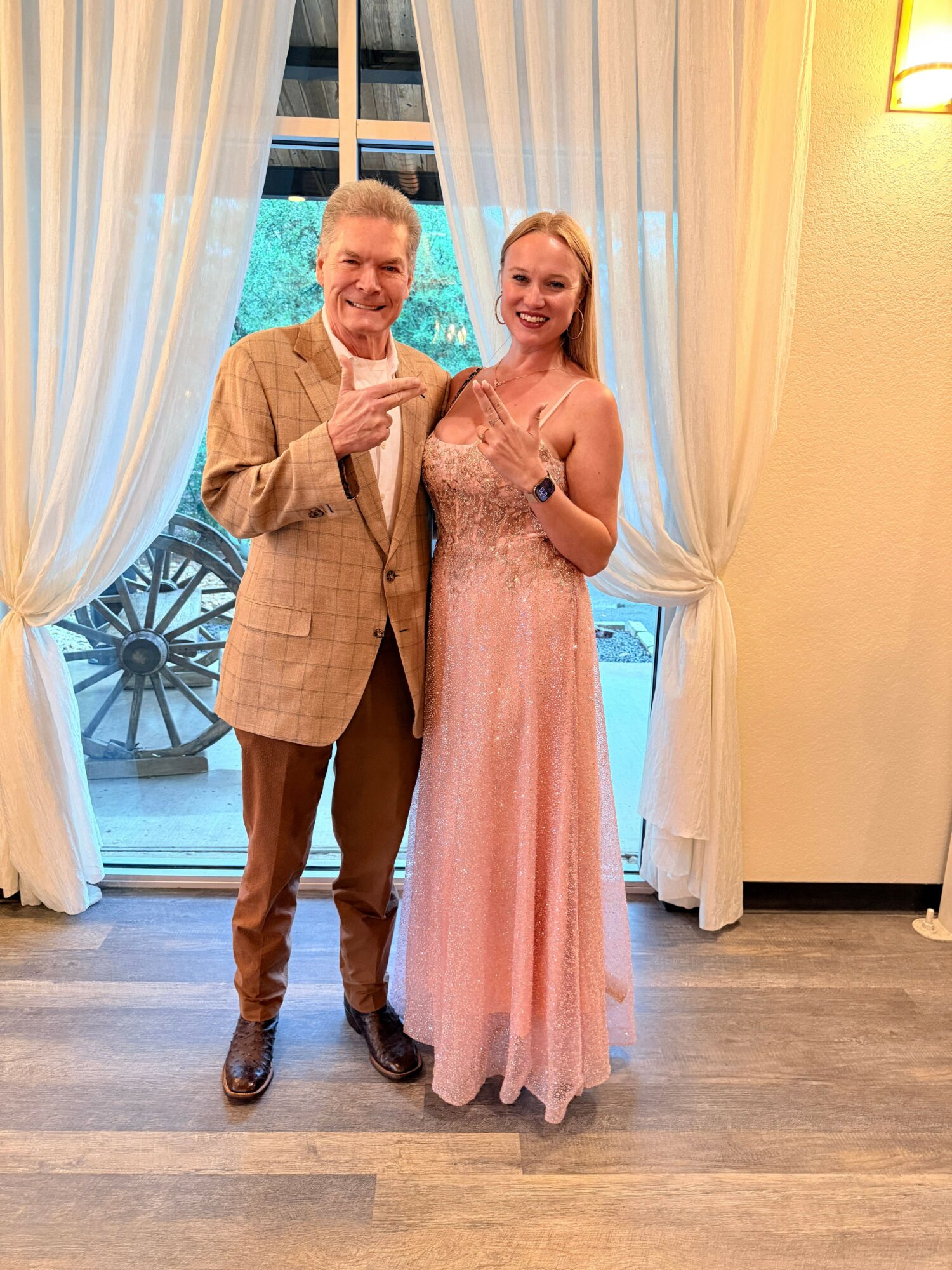 Two people stand indoors near a window with white curtains, dressed formally, smiling at the camera.