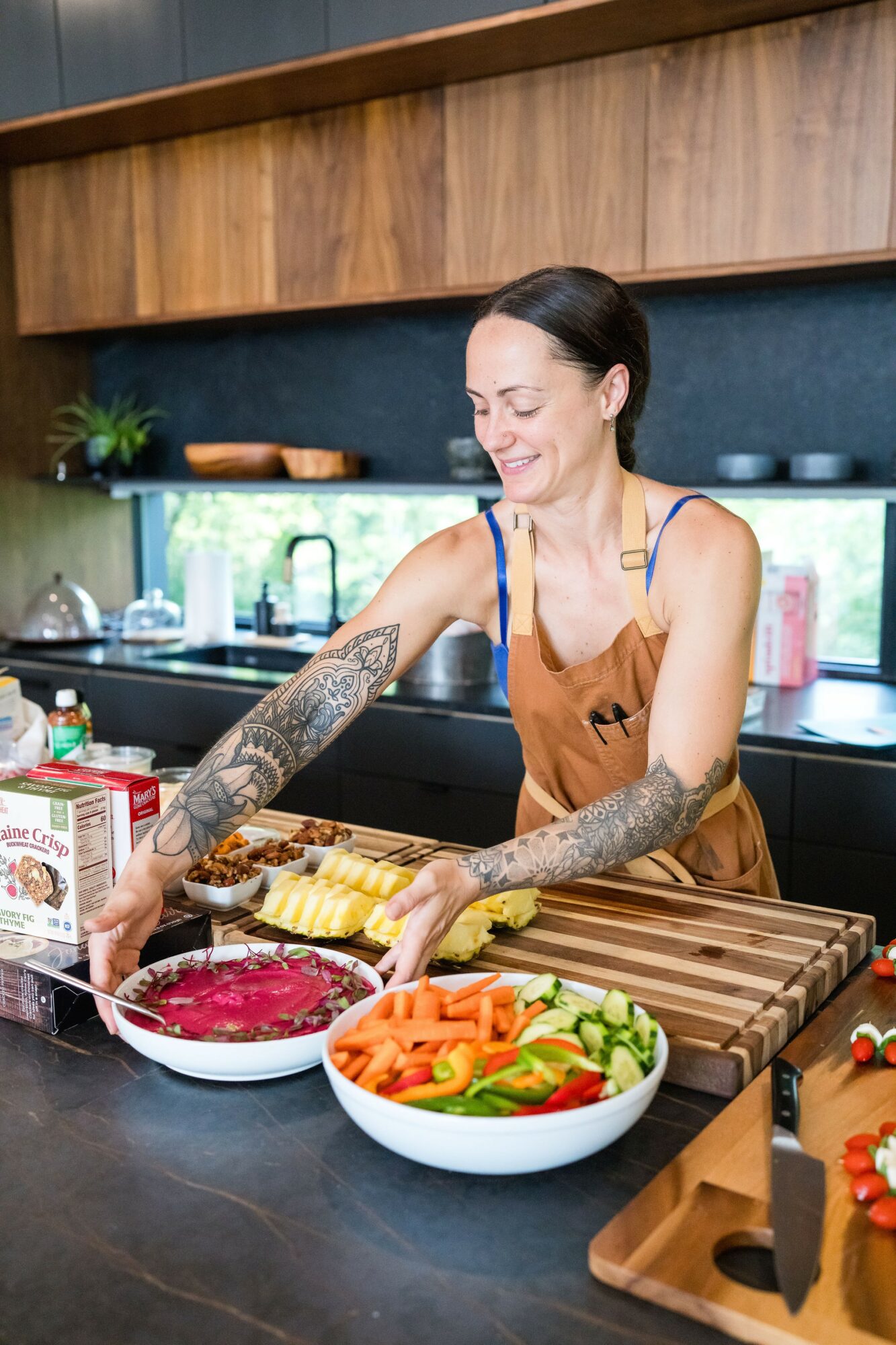 Woman with tattoos preparing food in a kitchen, smiling, with bowls of vegetables and ingredients on the counter.