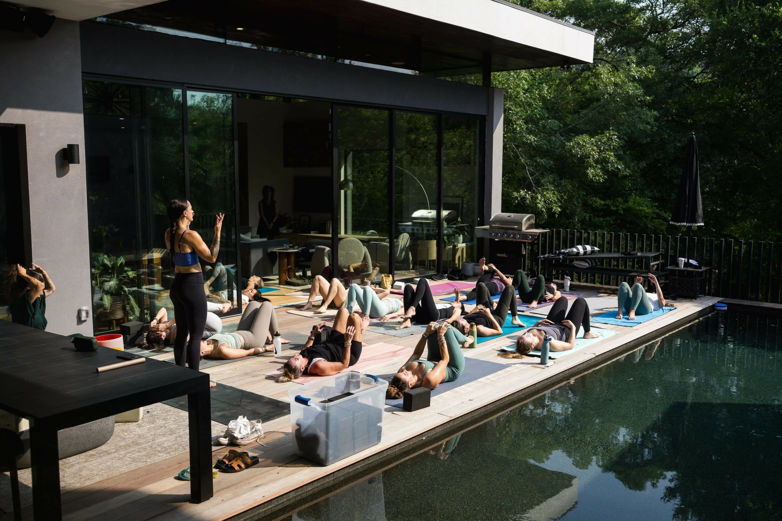 Group of people participating in an outdoor fitness class by a pool, instructor leading exercises, sunny day, modern house in background.