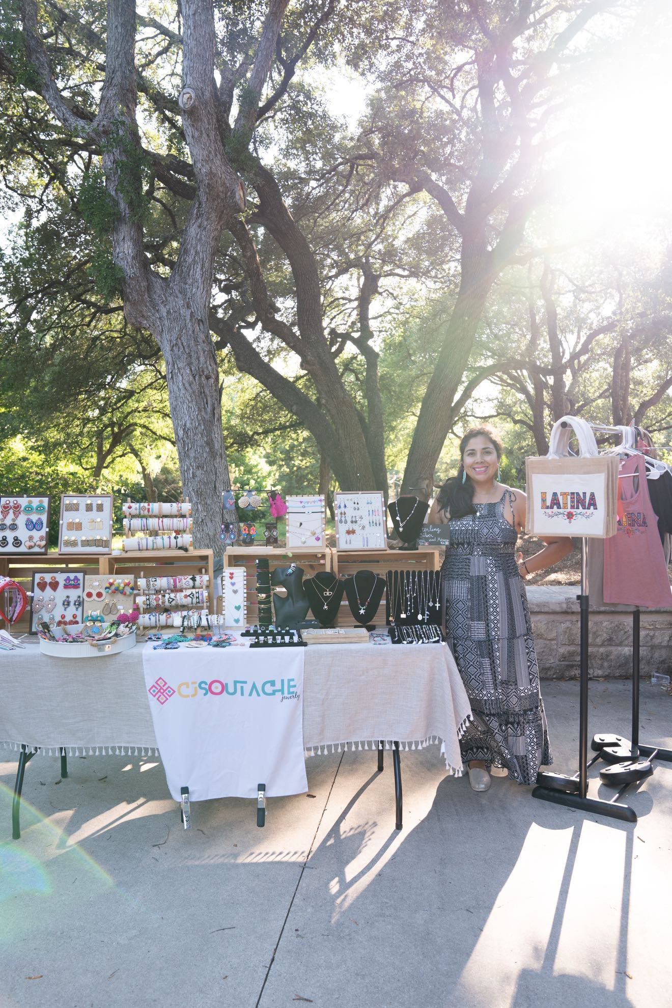 Person standing next to a table with handmade items outdoors under trees, sunlight shining brightly.