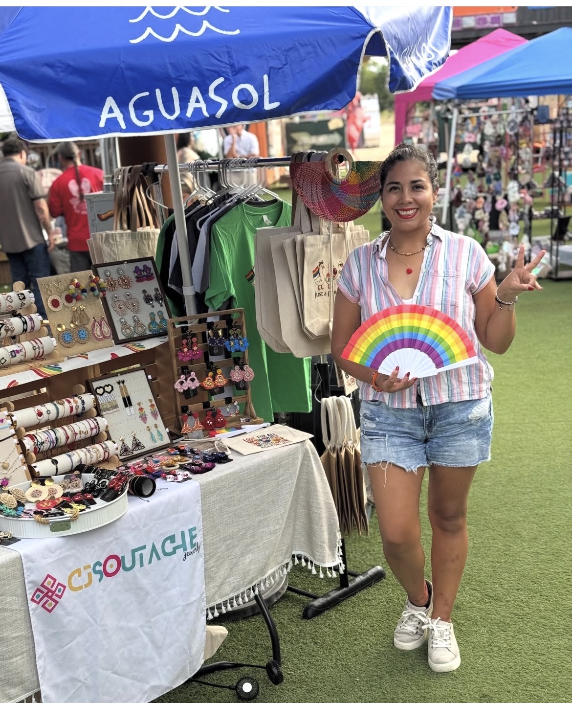 Young woman smiling holding a rainbow-shaped item at outdoor market stall with accessories and clothing, blue canopy overhead.