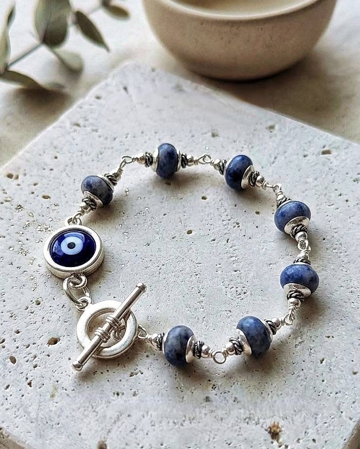 Blue and silver beaded bracelet with a toggle clasp on a textured white surface, with a bowl and plant in background.