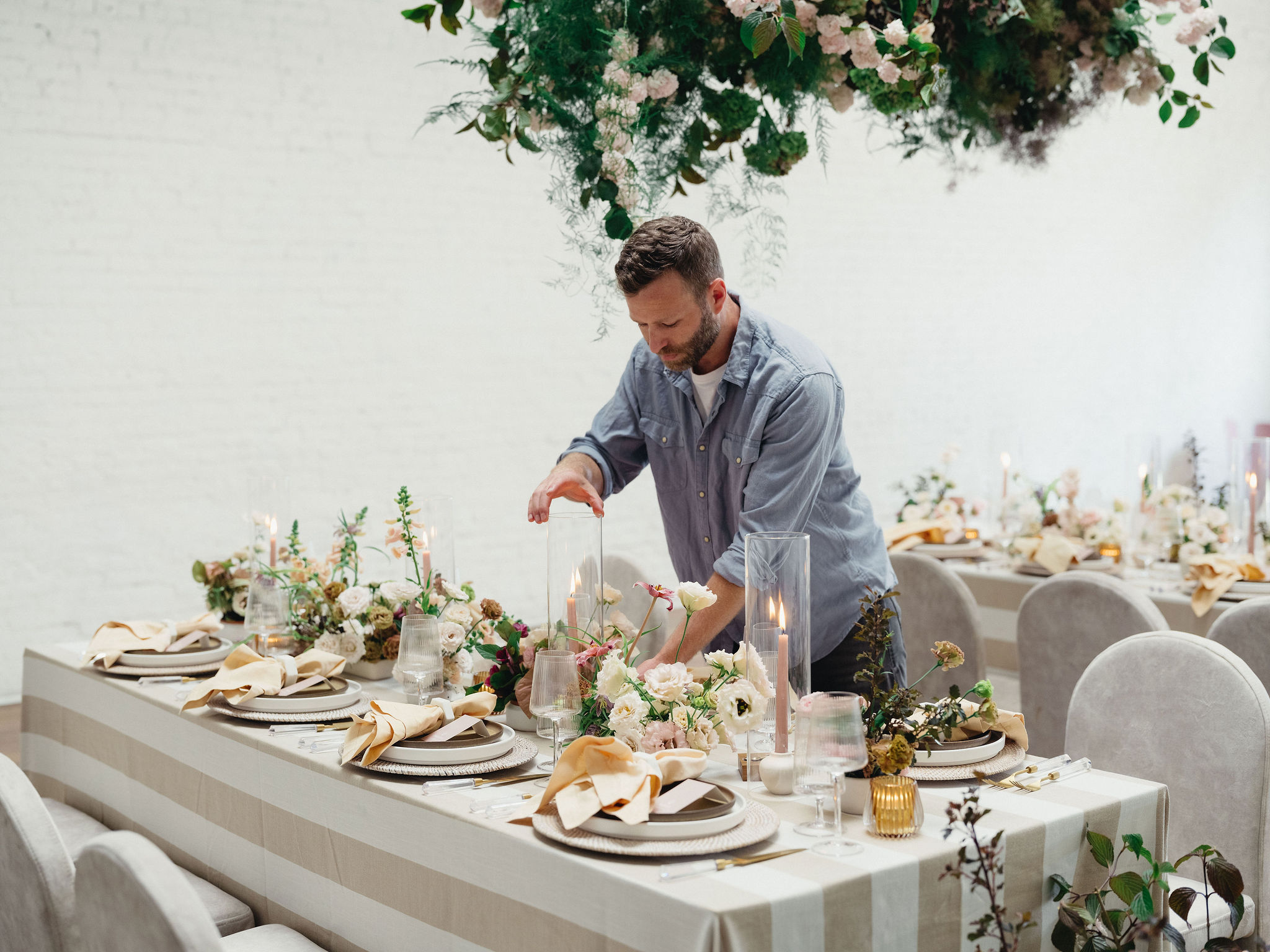 Man arranging a decorated table with flowers and candles, set for a formal event, in a bright room.