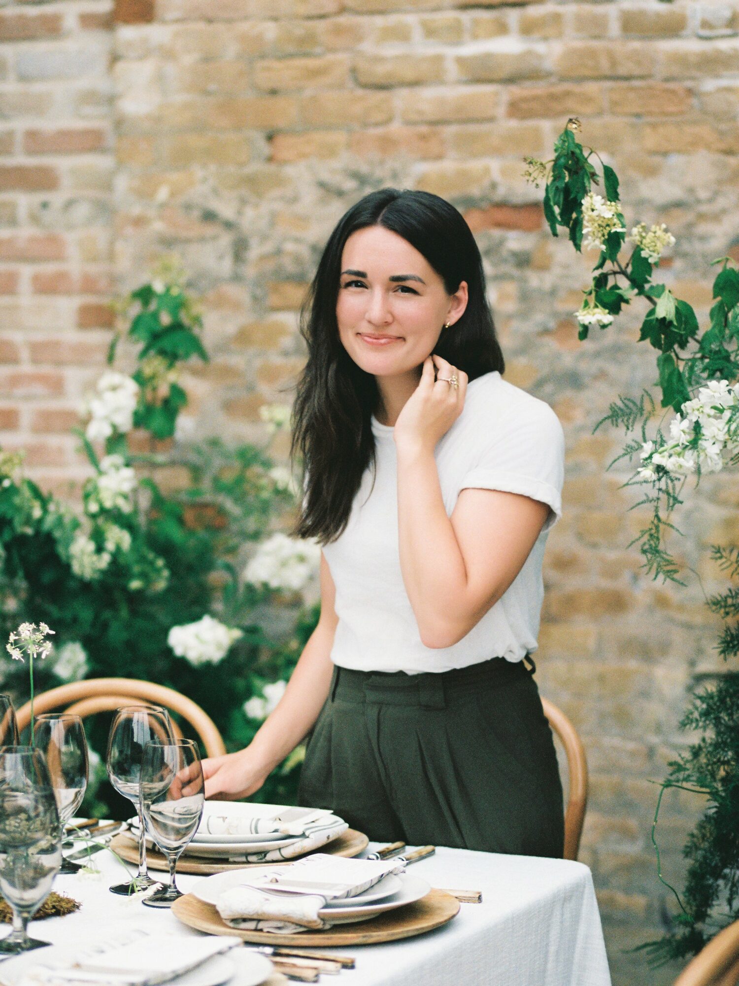 Young woman with dark hair in a white top standing beside a table outdoors, brick wall background, surrounded by green plants and white flowers.