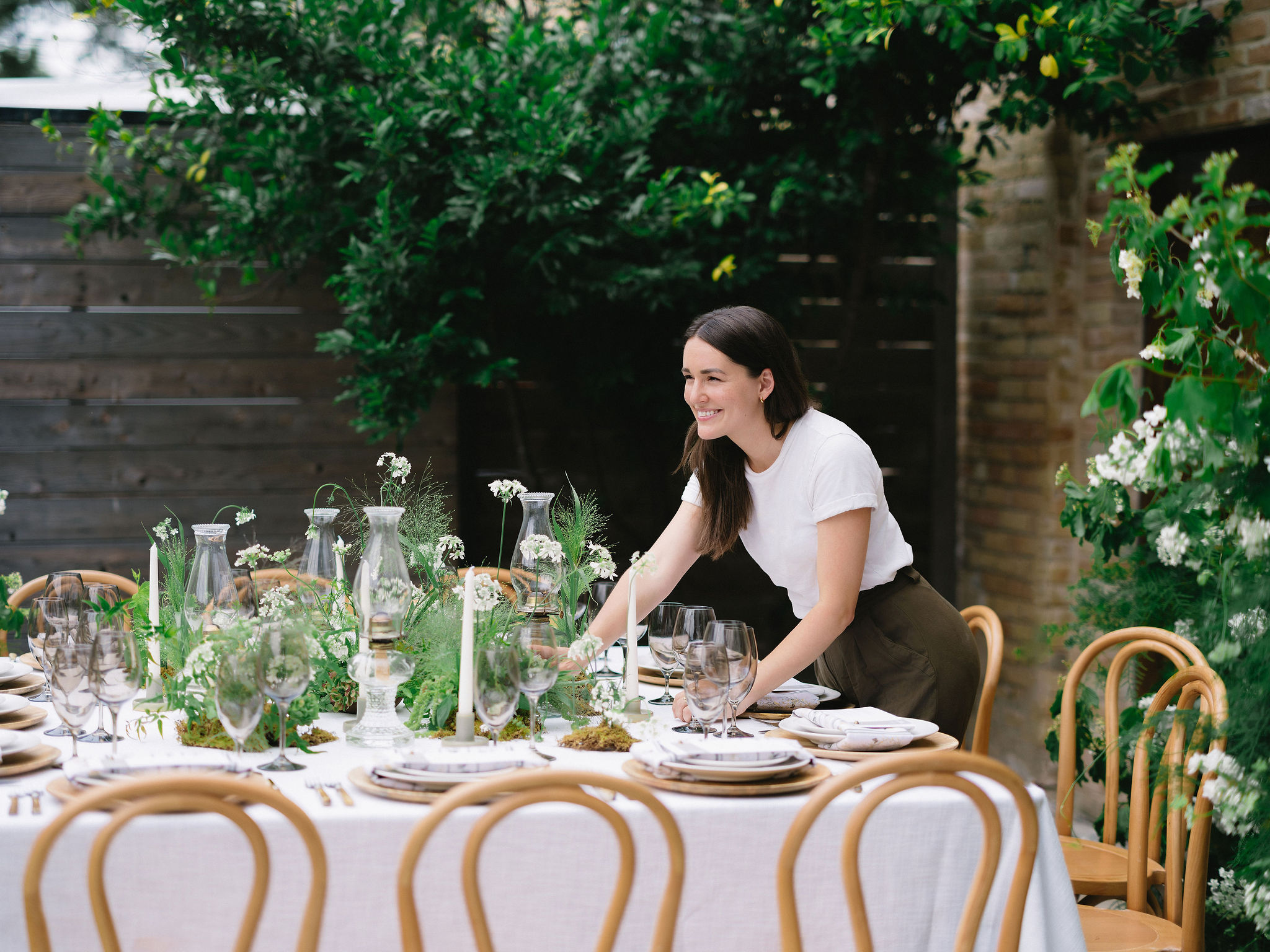 Woman setting a table outdoors with greenery and wooden fence in background.