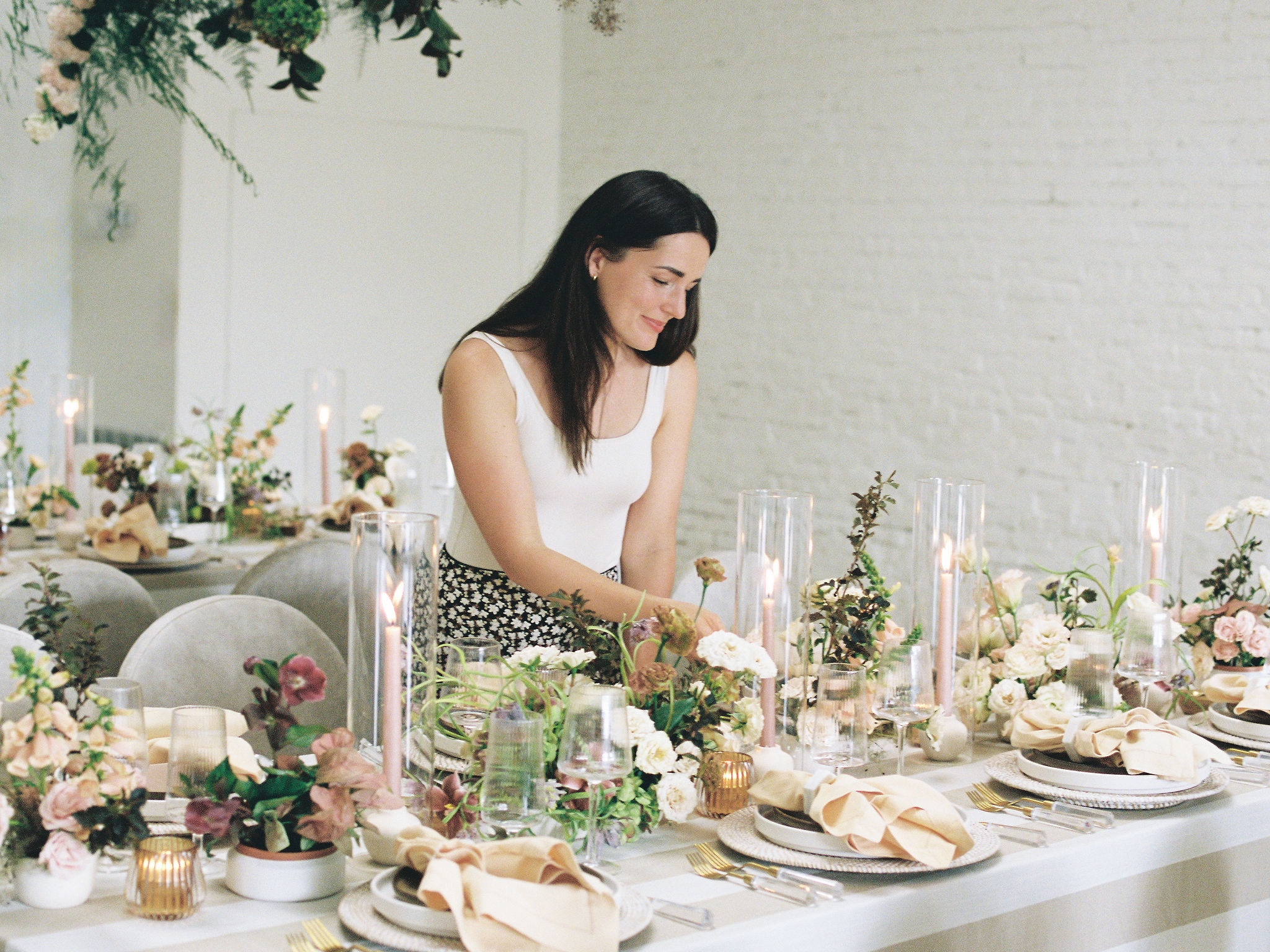 Woman arranging a table with floral centerpieces and candles, set for a formal event, in a bright room.