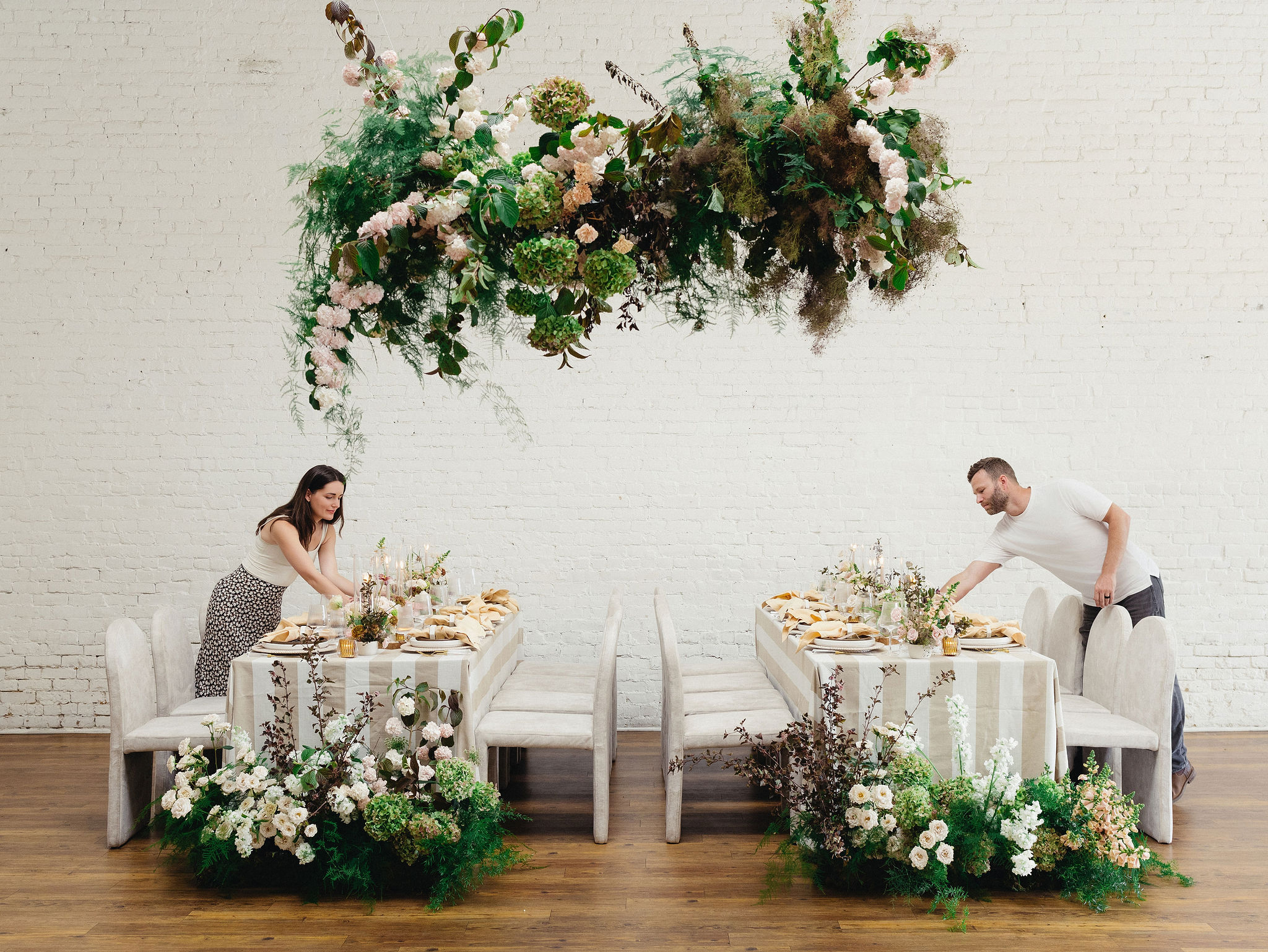 Two people setting tables with floral arrangements, hanging floral decoration above, in a bright room with white brick wall.