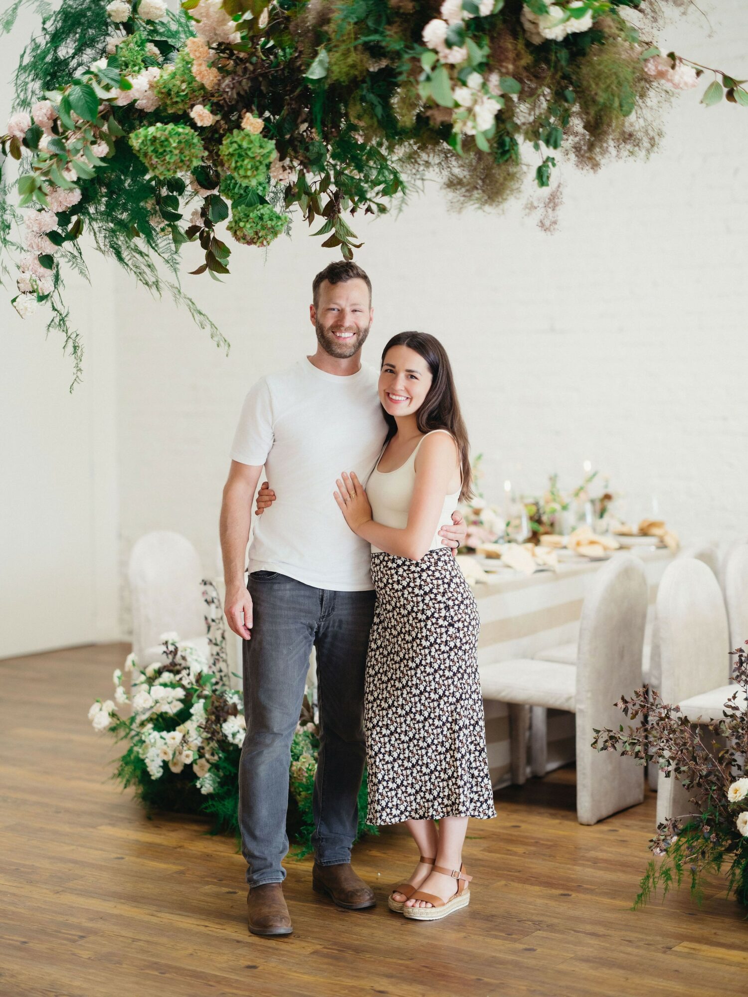 A man and woman stand together indoors, smiling, with a decorated table and floral arrangements in the background.