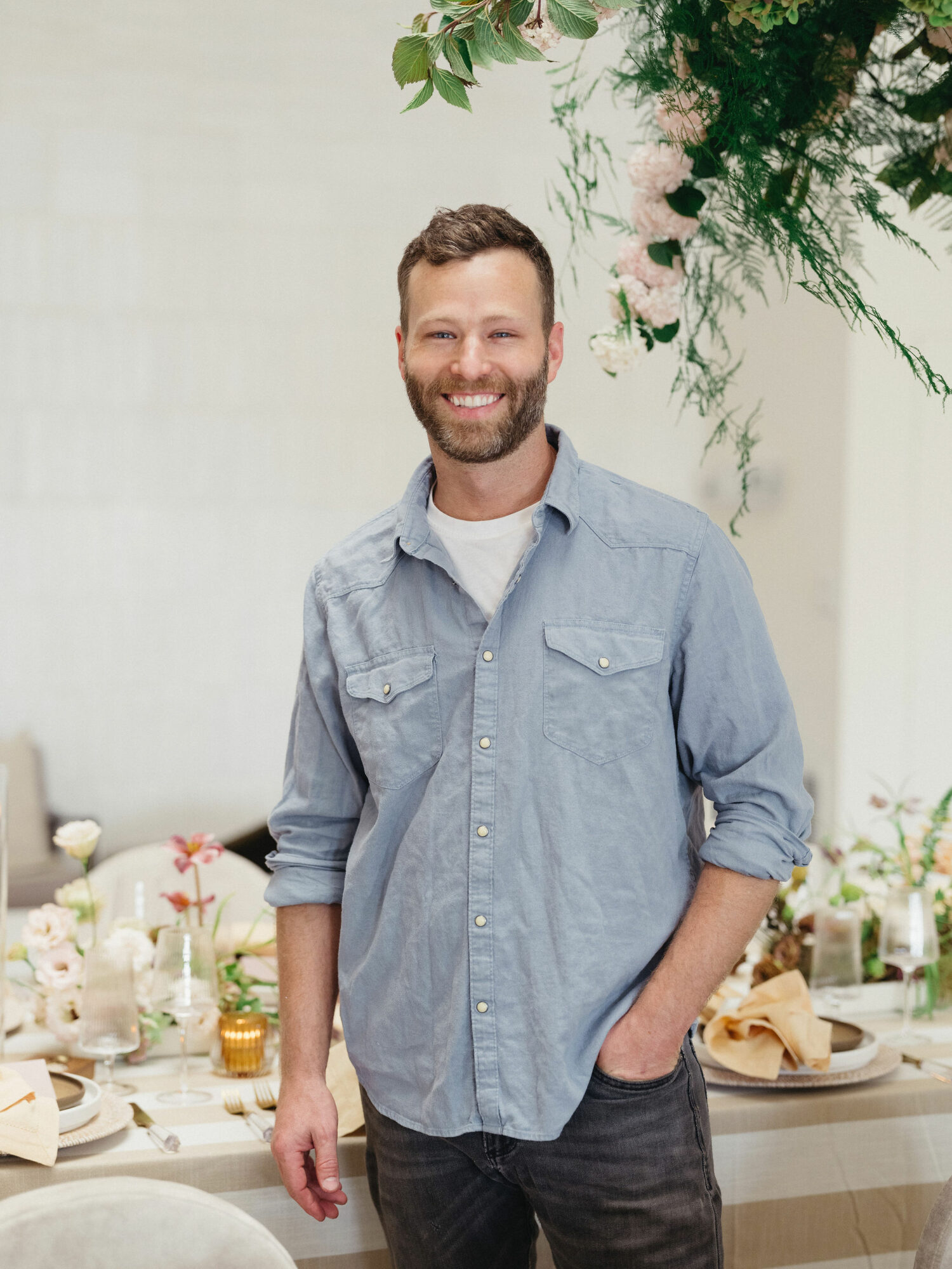 Smiling man with beard in a light blue shirt standing in a decorated indoor setting.